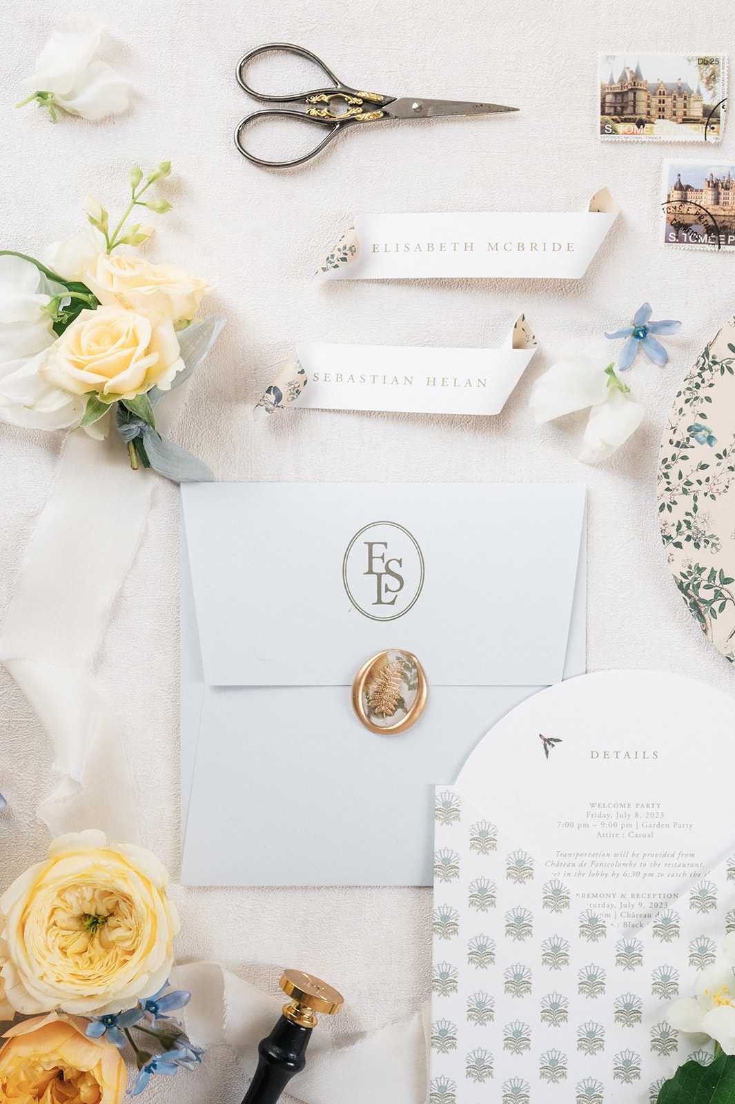 A flat lay detail shot of a wedding stationery suite arranged on a white textured surface. The suite includes a pale blue envelope sealed with a gold wax seal featuring a botanical fern motif and monogrammed with the initials 'ESL' in an oval crest, two white place cards reading 'Elisabeth McBride' and 'Sebastian Helan' with delicate floral detailing, a details card with a sage green block-print botanical pattern listing event information including a welcome party on Friday July 8, 2023 and ceremony and reception on Saturday July 9, 2023 at Château de Fonscolombe, and a floral-patterned envelope liner. Decorative postage stamps featuring a French chateau are placed in the upper right corner, alongside ornate antique-style scissors with gold-accented handles and a black and gold wax seal stamp. Soft yellow garden roses, white lisianthus, and small blue delphinium blooms are scattered around the edges of the arrangement, along with sheer white ribbon, completing the classic French-inspired stationery flat lay.