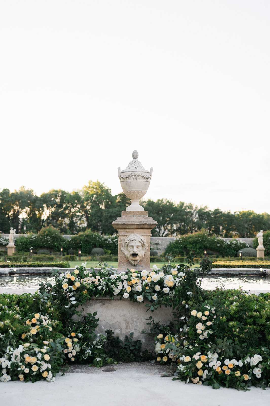 A detail shot of a formal French garden featuring a carved stone fountain pedestal topped with a decorative urn, adorned with a grotesque face mask spout at its base. The pedestal is surrounded by a lush ground-level floral arrangement composed of white and soft yellow roses with dark green foliage, arranged in sweeping clusters that frame the base of the structure and extend outward on both sides. A long rectangular reflecting pool sits directly behind the fountain, with manicured hedgerows, classical marble statues, and structured garden beds visible in the background. The overall decor palette is white, cream, and butter yellow against deep green, consistent with a classic French formal garden aesthetic. Potential venue feature image.