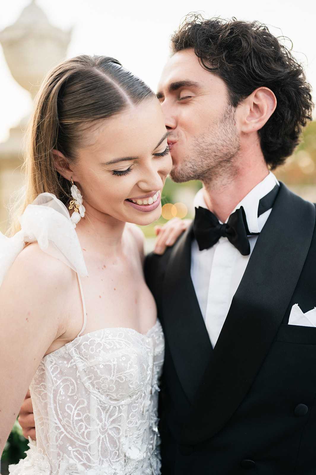 A close-up couples portrait of a bride and groom during what appears to be an outdoor photo session, with a softly blurred background showing warm golden bokeh. The groom, wearing a black tuxedo with satin lapels, black bow tie, and white pocket square, leans in to kiss the bride on her cheek. The bride wears a white strapless gown with intricate beaded and sequined floral embroidery, a sheer organza shoulder bow detail, and gold floral drop earrings; her hair is pulled back sleekly with a center part. The styling is classic and formal, with a polished, modern finish.