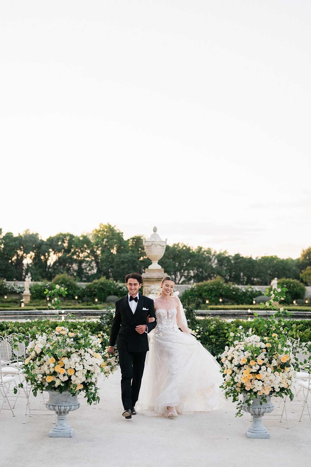 A bride and groom walk arm-in-arm through a formal outdoor garden setting at dusk, photographed in a wide portrait orientation. The groom wears a classic black tuxedo with a bow tie and white pocket square, while the bride wears a strapless, corseted ballgown with a voluminous tulle skirt and delicate lace detailing, lifting the skirt slightly as she walks. Flanking the couple are two large stone urns overflowing with garden roses in white, cream, and soft yellow, accented with trailing greenery, alongside white wrought-iron chairs arranged for a ceremony. The background features a formal French parterre garden with manicured hedgerows, small candle lights along the borders, classical stone urns on pedestals, and sculpted statuary, consistent with a historic chateau or palace garden. The overall styling is classic and formal, with a white and soft yellow floral palette. Potential venue feature image.