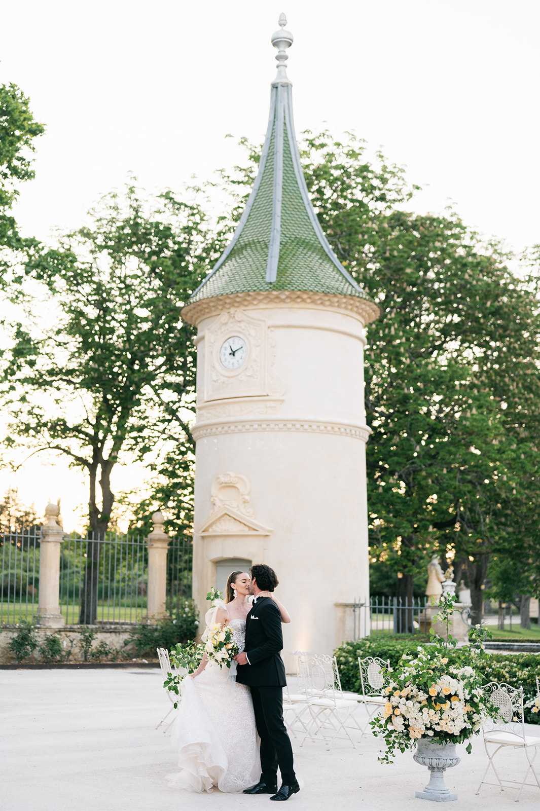 A bride and groom share a kiss outdoors in front of a cream-colored chateau turret with an ornate green tiled conical roof and a clock face, set within a formal garden courtyard. The bride wears a full-length white ballgown with a strapless neckline and carries a loose, trailing bouquet of white blooms and trailing greenery with soft yellow accents, while the groom wears a classic black tuxedo. White wrought-iron garden chairs are arranged nearby, and a large white stone urn overflows with an arrangement of ivory and soft yellow roses mixed with lush greenery. The wide portrait shot captures the couple centrally framed against the architectural tower, with warm evening light filtering through the surrounding trees. Potential venue feature image.