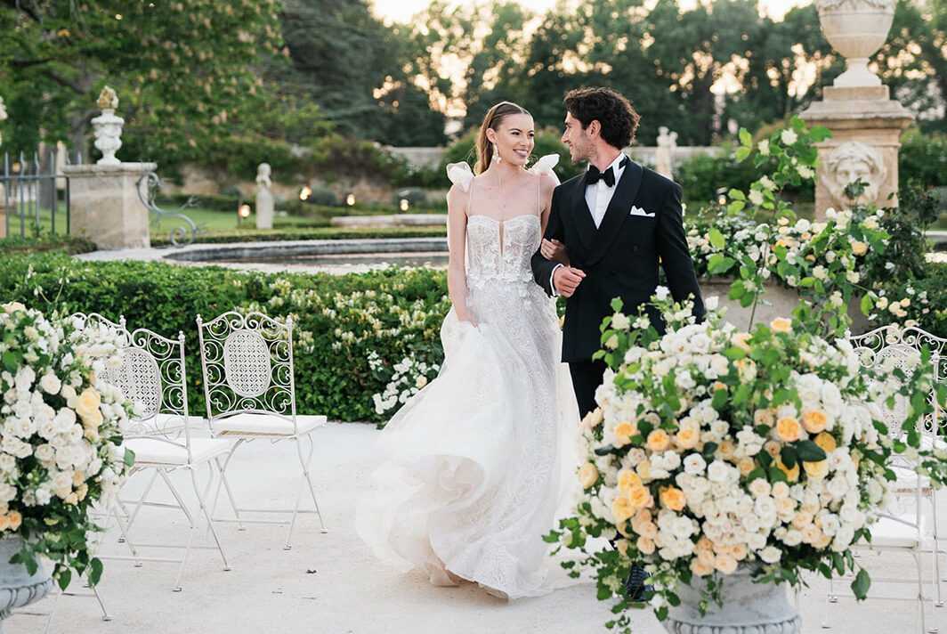 A couple walks arm-in-arm through an outdoor formal garden ceremony space at golden hour, smiling at each other. The bride wears a strapless ball gown with a 3D floral lace bodice, oversized bow shoulder details, and a full tulle skirt, while the groom wears a black tuxedo with bow tie and white pocket square. Large stone urns on either side overflow with arrangements of white and soft peach/yellow garden roses and trailing greenery, and white wrought-iron bistro chairs line the aisle. The formal garden setting features manicured box hedging, ornamental stone balustrades with urn finials, and small candle lights visible in the background. This is a mid-distance portrait shot capturing the full length of both subjects with the floral installations framing the foreground.