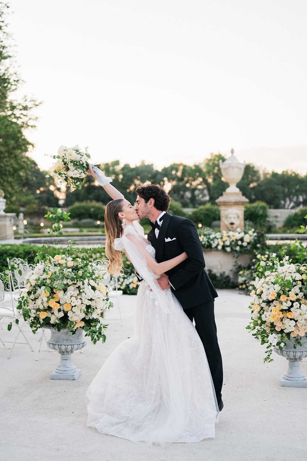 A couple shares a dip kiss during an outdoor ceremony or post-ceremony portrait in a formal French garden setting, with the bride raising her bouquet overhead with one arm. The bride wears a full-skirted white lace ball gown with sheer long gloves and drop earrings, and carries a loose bouquet of ivory and soft yellow roses with greenery. The groom is dressed in a classic black tuxedo with a white pocket square and black bow tie. Large stone urns on either side are overflowing with abundant arrangements of white and pale peach-yellow roses mixed with trailing greenery, establishing a cream and soft yellow floral palette throughout. The formal garden backdrop features manicured hedges, a stone lion-head fountain wall feature, and white folding chairs visible to the left. The shot is a full-length portrait taken at dusk in warm, fading natural light.