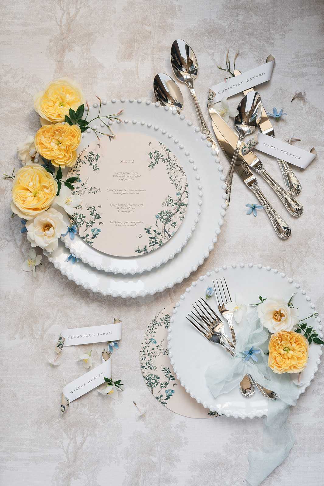 A flat lay detail shot of a wedding reception place setting styled on a neutral grey-white textured linen surface. The main place setting features two stacked white bobble-edge charger plates topped with a circular printed menu card decorated with a botanical illustration in soft sage green and dusty blue, with a blush background. Scattered around the plates are yellow garden roses, white roses, and small dusty blue flowers. Silver ornate cutlery — two spoons, a knife, and butter knife — is arranged to the right alongside two white ribbon place cards reading 'Christian Banero' and 'Maria Spenser.' A second partial place setting is visible at the bottom of the frame, with a white bobble-edge plate holding silver forks wrapped in a pale ice-blue linen napkin tied with a matching ribbon, topped with a yellow garden rose and white florals. Additional place cards reading 'Veronique Saban' and 'Marcus Hursen' are displayed at lower left, wrapped in the same botanical-print paper used on the menu card. The overall palette is soft yellow, ice blue, white, and sage green, with a classic yet delicate styling aesthetic.