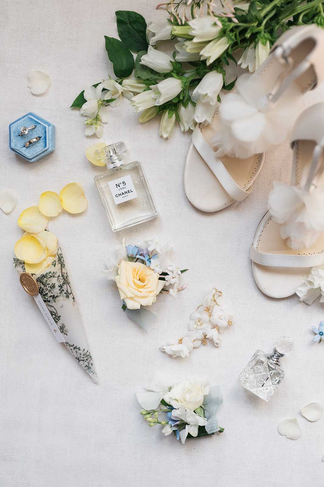 A flat lay detail shot of bridal accessories arranged on a light ivory surface, scattered with yellow and white rose petals. Visible items include a pair of white strappy heeled sandals, a bouquet of white tulips and lisianthus with green foliage, a blue hexagonal velvet ring box holding two wedding bands, a bottle of Chanel No. 5 L'Eau perfume, a small crystal perfume bottle, two boutonnieres featuring cream roses, white stephanotis, and pale blue flowers, a pair of delicate gold-and-white floral drop earrings, and a cone-shaped paper confetti holder with a botanical green-and-white print sealed with a gold wax stamp. The overall color palette is white, cream, pale yellow, and soft blue, reflecting a classic and refined styling theme.