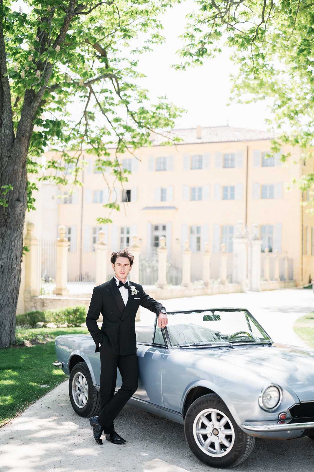 A groom portrait taken outdoors on the driveway of a French chateau, with the pale yellow facade of the manor house softly out of focus in the background. The groom is dressed in a black double-breasted tuxedo with a white dress shirt, black bow tie, white pocket square, and a small white floral boutonniere, leaning casually against a vintage powder blue convertible sports car. The shot is a full-length portrait with a classic, formal styling aesthetic. Potential venue feature image.