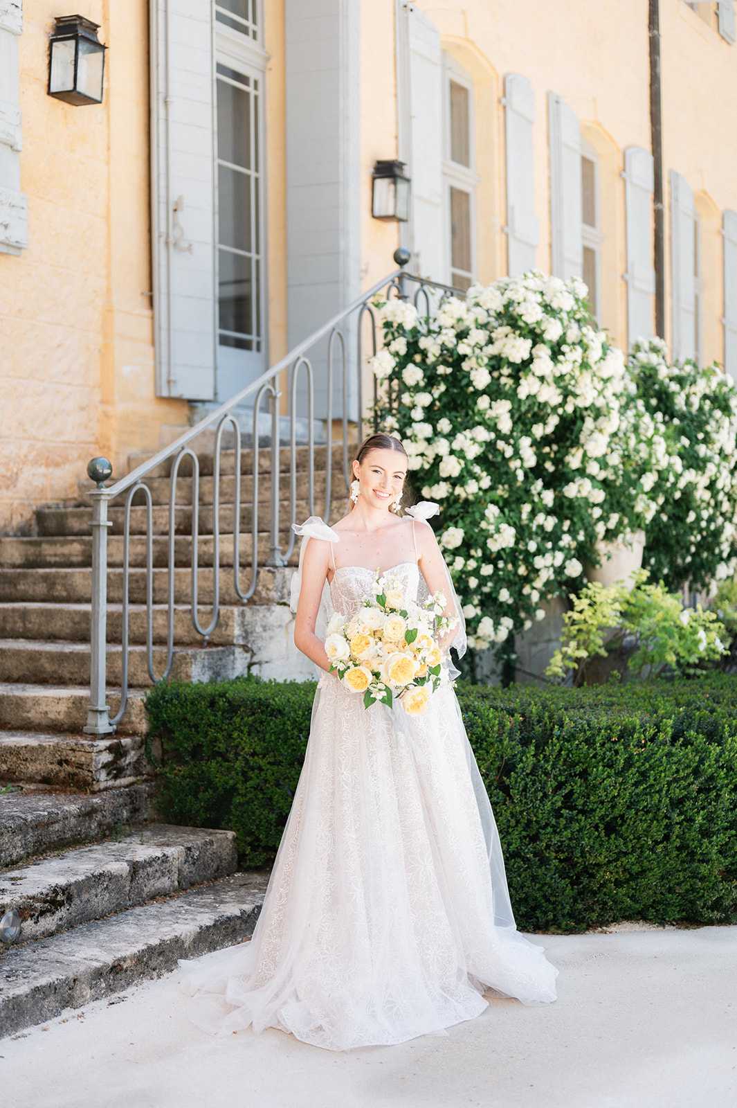 A bridal portrait taken outdoors in front of a French chateau with warm yellow stone facade, pale grey shutters, and stone steps with wrought-iron railings. The bride stands alone on a gravel courtyard, wearing a full A-line lace gown with a sweetheart neckline and white bow shoulder straps, holding a bouquet of soft yellow garden roses and white blooms with green foliage. Her hair is pulled back and she wears white floral drop earrings. The styling is classic and refined with a soft yellow and white floral palette. The shot is a full-length portrait with the chateau entrance staircase and manicured boxwood hedges visible in the background. Potential venue feature image.