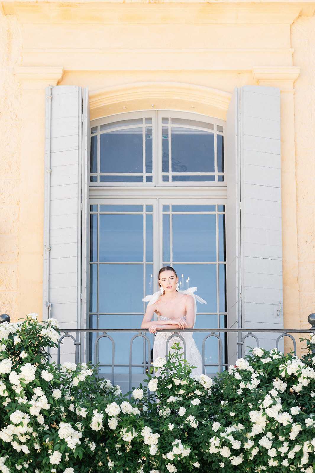 A bridal portrait of a single bride standing on a wrought-iron balcony of a classic French chateau with warm yellow limestone façade and pale grey shutters flanking an arched window. The bride wears a deep V-neck, largely backless lace gown with a white feather or bow shoulder accent, and large statement earrings, with her hair pulled back sleekly. She leans gently on the balcony railing, looking directly at the camera. The foreground is filled with dense clusters of white roses in full bloom lining the base of the balcony, creating a natural floral frame. The overall styling is classic and refined, with a white-and-grey palette. Medium-distance portrait shot framed symmetrically by the architecture. Potential venue feature image.