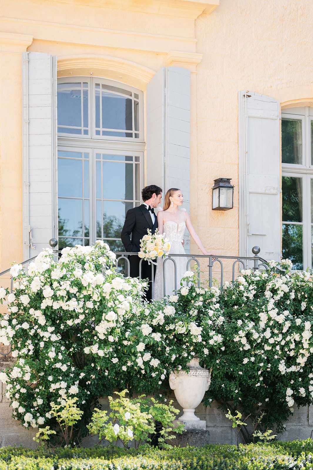 A couple portrait taken outdoors on the terrace balcony of a French chateau, shot from below as a wide portrait. The groom wears a black tuxedo with a bow tie, and the bride wears a fitted strapless lace gown with a sheer corset bodice; she holds a bouquet of white and soft yellow blooms. The pair stand close together at a wrought-iron railing, with the warm ochre-yellow facade of the chateau and pale grey shuttered arched windows directly behind them. The foreground is filled with densely flowering white rose bushes spilling over a stone balustrade, flanked by a classical stone urn planter, creating a natural frame around the couple. The overall styling is classic and formal with a white and green floral palette. Potential venue feature image.