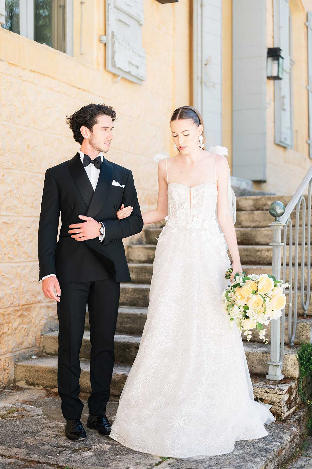 A couple portrait taken outdoors on stone steps alongside a French chateau or manor house with warm yellow rendered walls and pale blue-grey shutters. The groom wears a black double-breasted tuxedo with a bow tie and white pocket square, while the bride wears a fitted ivory A-line gown with a sweetheart neckline, thin spaghetti straps with small floral shoulder details, and an embroidered or lace-patterned skirt. She carries a loose bouquet of yellow garden roses, white blooms, and green foliage, and wears statement floral drop earrings with her hair pulled back sleekly. The groom looks toward the bride while she glances downward, her hand resting on his arm. The shot is a medium full-length portrait with soft warm golden-hour light, styled in a classic, formal aesthetic.