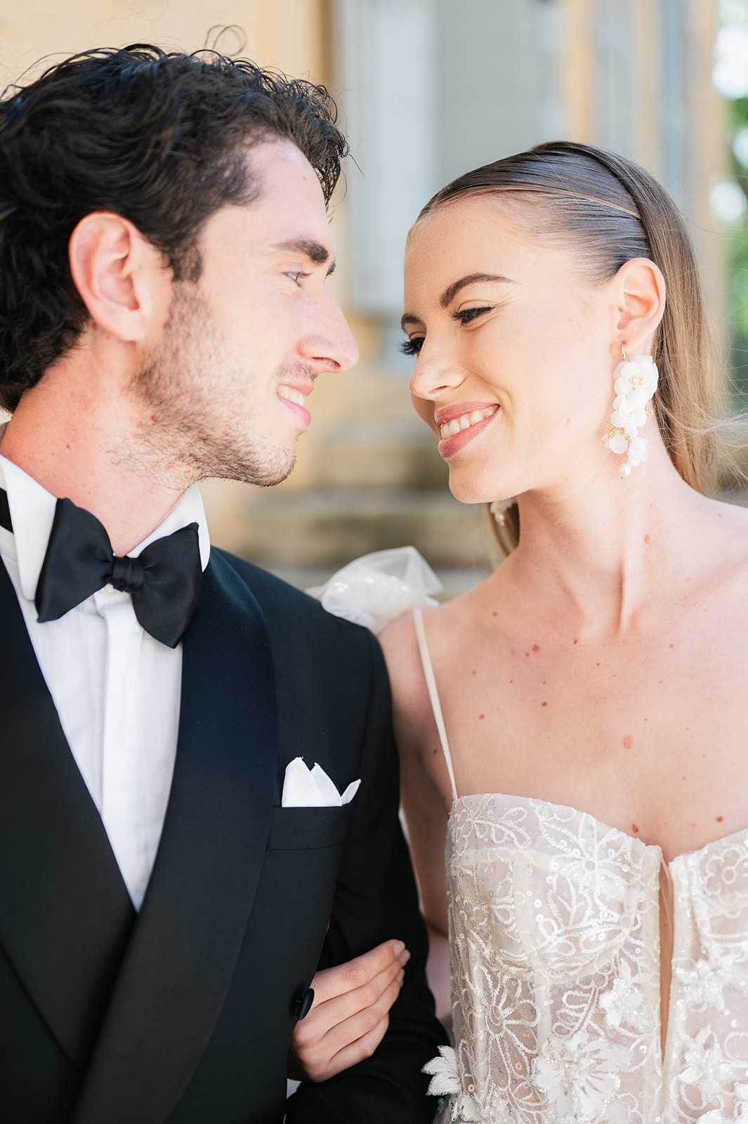 A close-up portrait of a bride and groom looking at each other and smiling outdoors, with a blurred architectural backdrop. The groom wears a black tuxedo with a black bow tie, white dress shirt, and a white pocket square. The bride wears an ivory lace and beaded spaghetti-strap gown with a sweetheart neckline, her hair slicked back sleekly, and she accessorizes with large white floral drop earrings with gold accents. The styling is classic and formal, with a sharp black-and-white contrast in the groom's attire against the bride's ivory look.