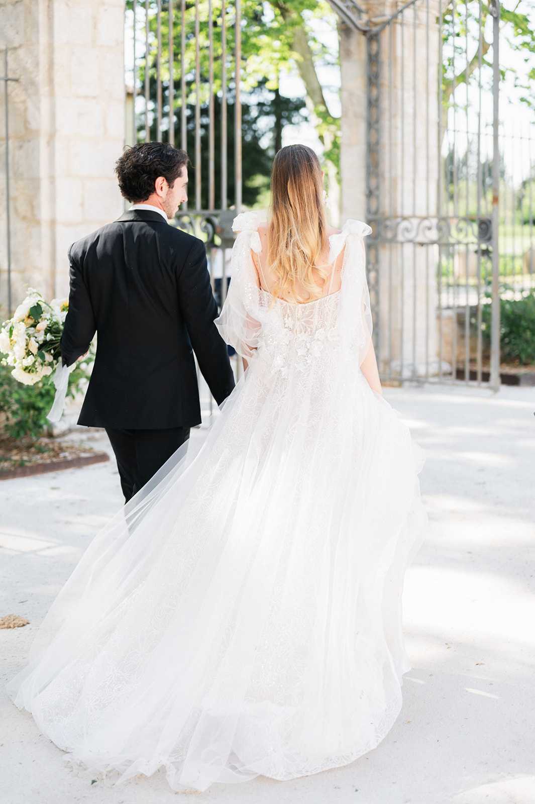 A couple walks hand-in-hand away from the camera toward a large ornate wrought-iron gate set between stone pillars, suggesting a French chateau or estate entrance. The groom wears a classic black suit and holds a bouquet of white and cream flowers with trailing ribbons, while the bride wears a white lace-bodiced gown with a voluminous tulle skirt, ruffled off-shoulder straps, and a long flowing train that fans out behind her. The bride's hair is loose and ombre-toned, worn down her back. The shot is a full-length portrait taken from behind, capturing the movement of the dress and the couple's forward motion in a natural, candid style.
