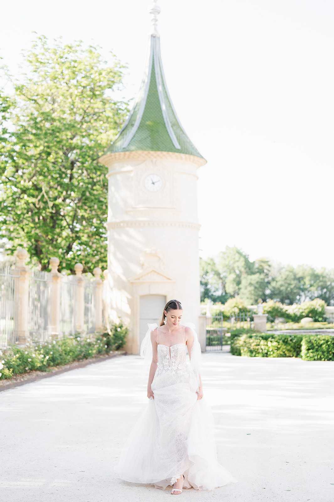 A bridal portrait taken outdoors on a gravel courtyard, with the bride standing alone looking downward. She wears a fitted ivory lace gown with spaghetti straps, a sweetheart neckline with floral appliqué bodice detail, and a flowing tulle skirt; she also wears drop earrings and strappy heeled sandals. In the background, slightly out of focus, stands a round chateau tower with a green pointed roof and a clock face, flanked by formal garden hedging, decorative stone balustrades, and iron gates. The composition is a full-length portrait shot with soft, bright natural light creating an airy, high-key feel. Potential venue feature image.
