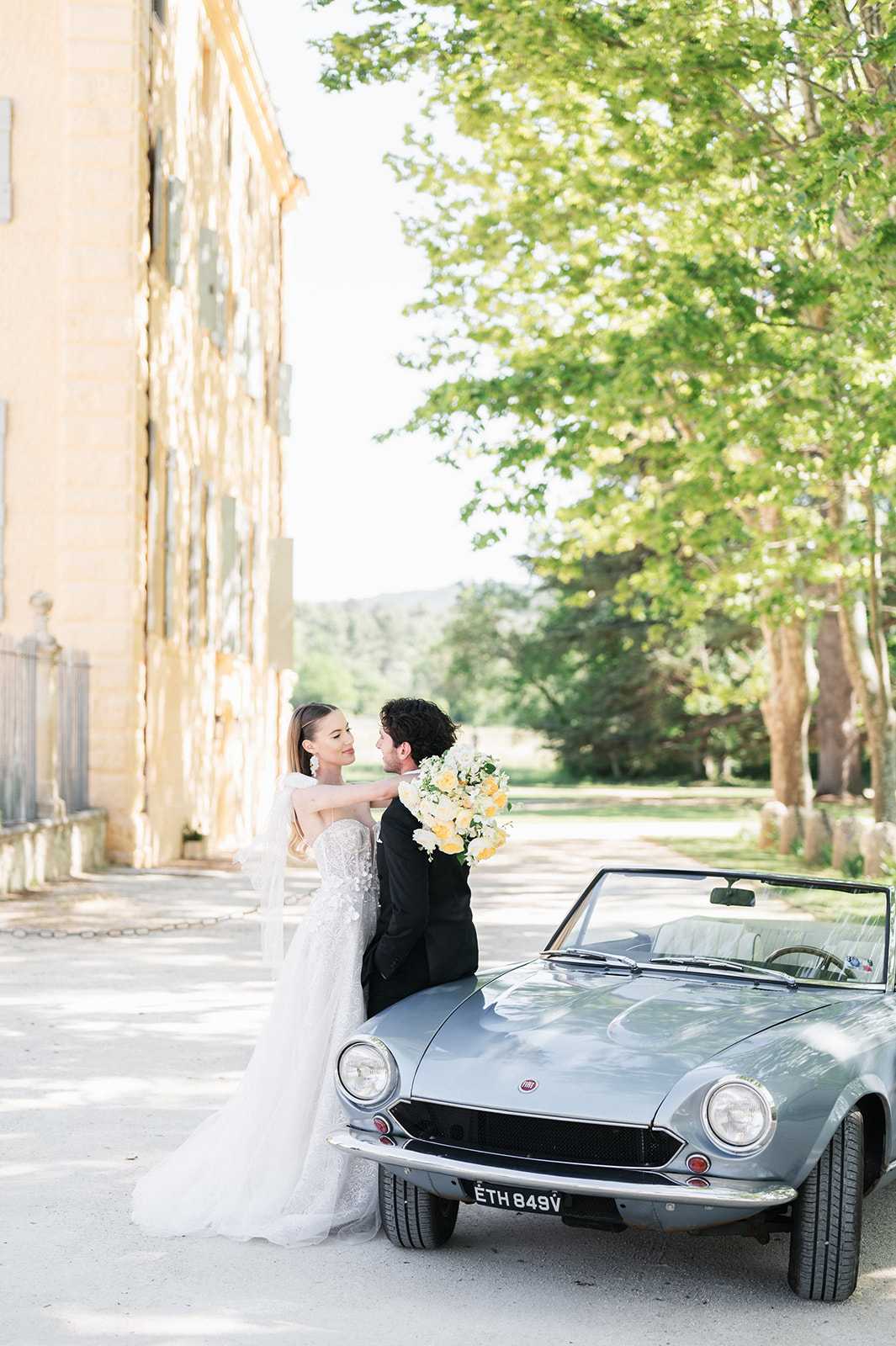 A couple portrait taken outdoors alongside the facade of a French chateau, with the bride and groom leaning against the hood of a vintage pale blue-grey Fiat convertible. The groom, dressed in a black tuxedo, sits on the front of the car while the bride stands beside him with her arm around his neck; they are gazing at each other. The bride wears a fitted, heavily embellished lace gown with a flowing tulle overlay and long veil, accessorized with statement drop earrings. She holds a loose, garden-style bouquet of soft yellow roses and white blooms with trailing greenery. The styling is classic and polished, combining the vintage car with the formal attire for a refined aesthetic. Medium portrait shot with the chateau's golden stone facade visible on the left.