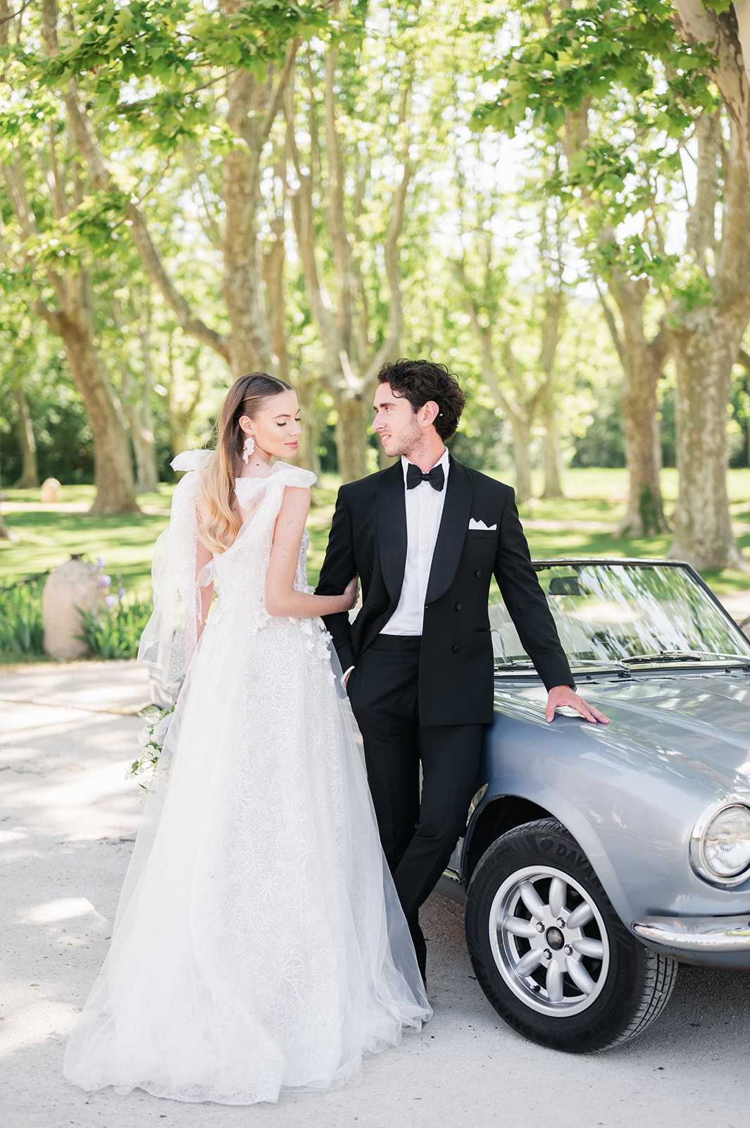 A couple portrait taken outdoors along a tree-lined driveway, with the bride and groom posed beside a vintage silver-grey convertible sports car. The bride wears a full A-line ivory lace gown with delicate 3D floral appliqué details, a large bow at the back shoulder, and a long flowing veil; she wears white drop earrings and her hair is worn loosely down. The groom wears a black double-breasted tuxedo with a black bow tie and white pocket square. The two are in close contact, with the groom resting his hand on the car and looking at the bride, while she looks slightly downward. The styling is classic and formal with a French countryside aesthetic. The shot is a medium full-length portrait with soft natural daylight filtering through the tree canopy.