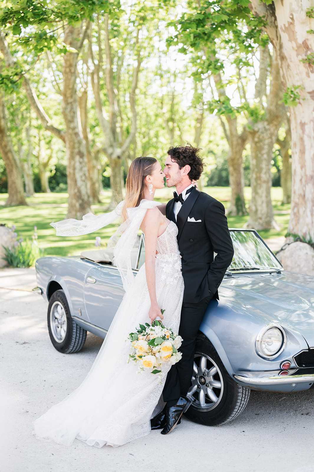 A couple portrait taken outdoors in a tree-lined driveway, with the groom leaning against a vintage pale blue convertible sports car while the bride leans in close as they are about to kiss. The bride wears a fitted, heavily embellished ivory lace gown with a flowing sheer cape or oversized bow detail at the shoulder, and holds a loose bouquet of soft yellow peonies, white blooms, and green foliage. The groom is dressed in a classic black tuxedo with a bow tie, white pocket square, and black patent leather shoes. The styling is classic and polished, with a French countryside aesthetic suggested by the plane tree-lined gravel drive in the background. The shot is a full-length portrait with the vintage car as a deliberate styling prop.