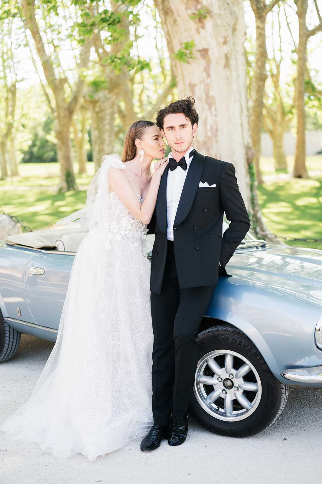 A couple portrait taken outdoors in a tree-lined driveway, with the bride and groom posed leaning against a vintage powder-blue convertible sports car. The bride wears a white strapless ball gown with all-over floral lace appliqué, a cathedral-length veil, and large white statement earrings, with her hair worn sleek and straight. The groom wears a black double-breasted tuxedo with a black bow tie, white pocket square, and black lace-up boots. The styling is modern and classic, with a high-contrast pairing of the white gown against the black tuxedo. The shot is a full-length couple portrait with a slightly compressed background, giving the vintage car and tree-lined setting equal visual weight alongside the subjects.