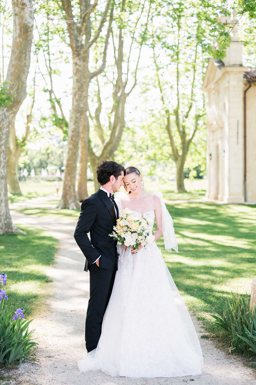 A couple portrait taken outdoors along a tree-lined gravel path, likely on the grounds of a French chateau or estate, with a stone classical building visible on the right edge of the frame. The groom, dressed in a black tuxedo with a bow tie, leans in close to the bride's cheek as she smiles downward. The bride wears a white ballgown with a fitted embellished bodice featuring thin spaghetti straps and a full tulle skirt, paired with a sheer veil draped over one arm and statement drop earrings. She holds a loose, garden-style bouquet composed of ivory roses, pale peach blooms, white stock flowers, and green foliage. The overall styling is classic and refined, with a neutral ivory and blush floral palette. The shot is a full-length portrait with soft natural midday light filtering through the plane tree canopy overhead.