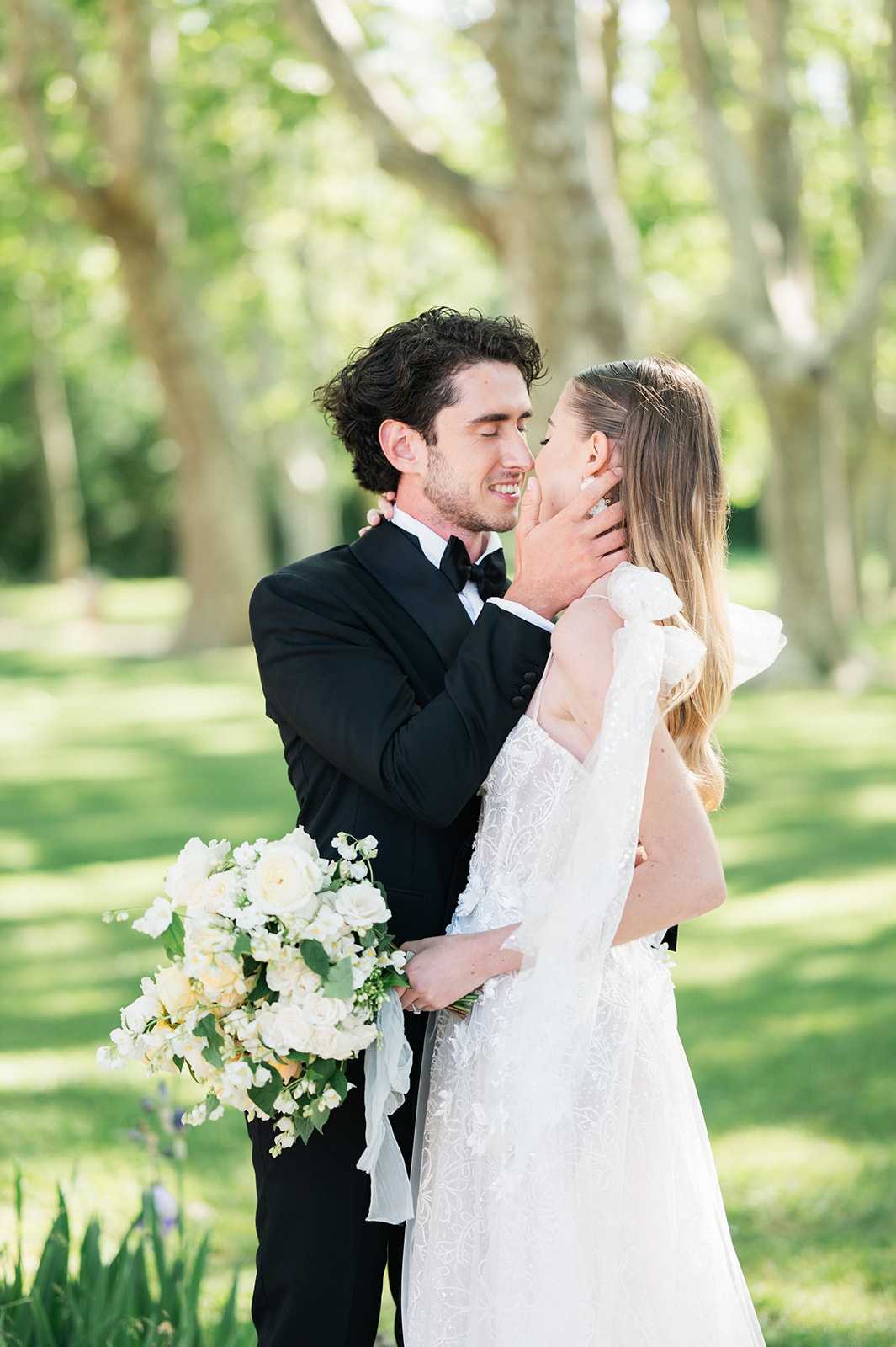 A couple portrait shot outdoors in a tree-lined park or garden setting, with the bride and groom leaning in to kiss. The groom wears a black tuxedo with a black bow tie, and the bride wears a white lace gown with delicate embroidery and distinctive oversized organza bow or ruffle shoulder details. She carries a large, loosely arranged bouquet of ivory garden roses, white sweet peas, lily of the valley, and greenery with trailing white ribbon streamers. The overall styling is classic and refined, with a white and ivory floral palette. Medium portrait composition with a shallow depth of field, placing focus on the couple against a softly blurred background of tall plane trees.