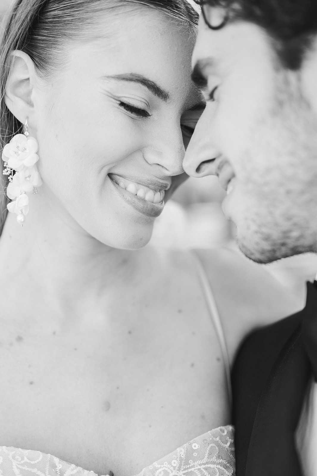 This is a black-and-white close-up portrait of a bride and groom sharing an intimate moment, their faces nearly touching as the bride smiles broadly. The image is shot in high contrast with bright highlights on the bride's face and soft shadows framing the groom, whose face is partially cropped at the right edge. The bride wears a strapless lace and sequin dress and distinctive drop earrings featuring floral and pearl details. The groom is wearing a dark suit jacket visible at the lower right corner of the frame.