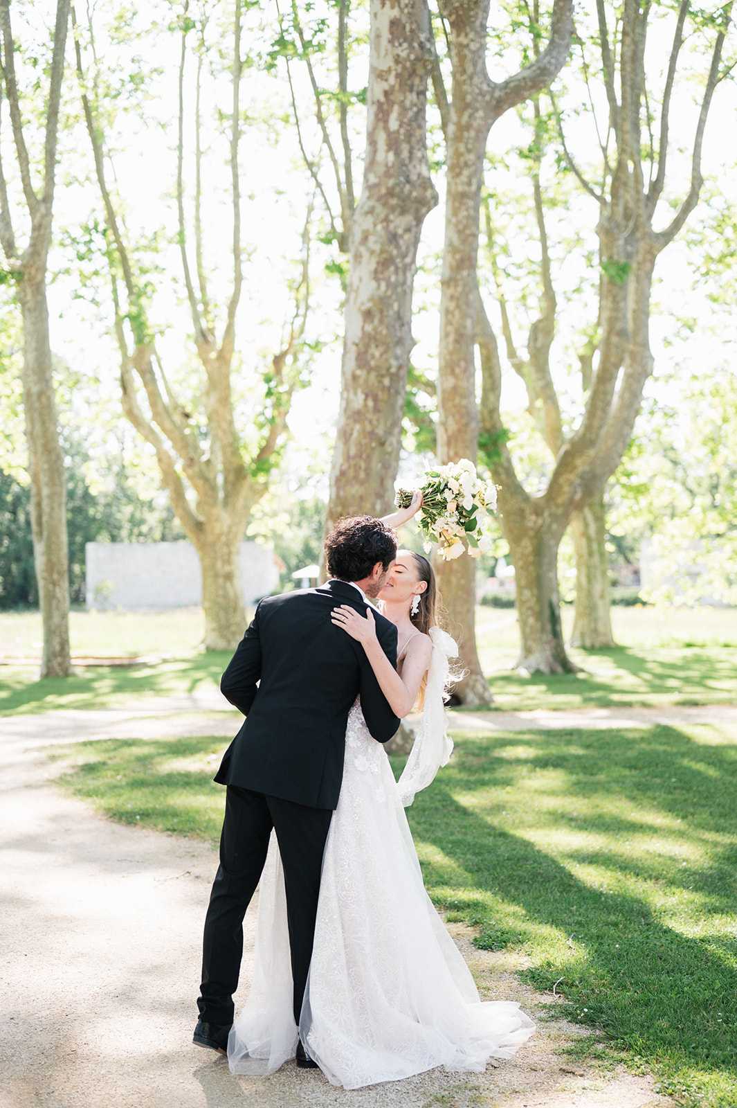 A couple shares a kiss during an outdoor portrait session along a gravel path lined with tall plane trees. The groom wears a classic black tuxedo, while the bride wears a fitted ivory lace gown with a flowing tulle overlay or cape detail; she holds a white and cream bouquet composed of large blooms with green foliage raised above her head. The styling is classic and clean, with a monochromatic black-and-white dress code contrasted against the bright, sunlit garden setting. The shot is a full-length portrait taken from behind and slightly to the side, capturing both figures from head to toe.