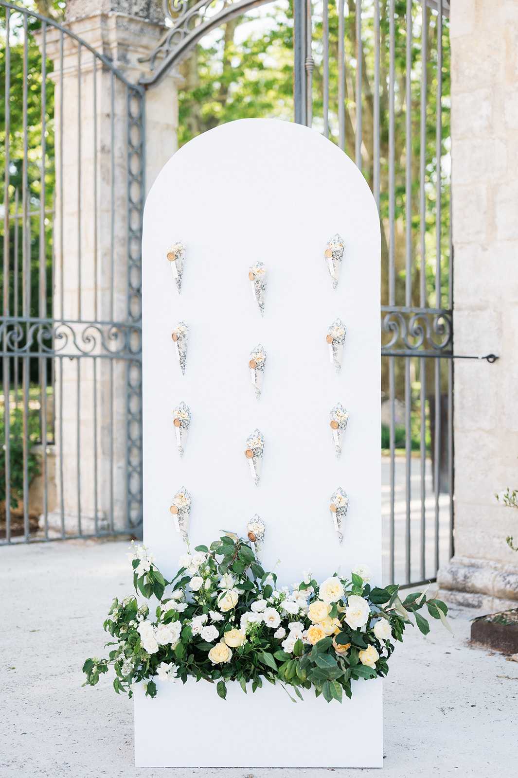 A wedding welcome or escort card display station positioned outdoors in front of ornate wrought iron gates at what appears to be a chateau or estate venue. The display consists of a tall white arch-shaped panel mounted on a white rectangular base, holding approximately twelve individual cone-shaped holders — each containing what appears to be a rolled paper escort card or favor with a small circular tag — arranged in a grid pattern across the panel. The base of the display is decorated with a lush arrangement of ivory and soft yellow garden roses, white ranunculus or similar blooms, and abundant green foliage including large leaves and trailing vines. The overall decor palette is white, ivory, pale yellow, and green, consistent with a classic or modern-classic wedding aesthetic. Wide shot, no people visible. Potential venue feature image.