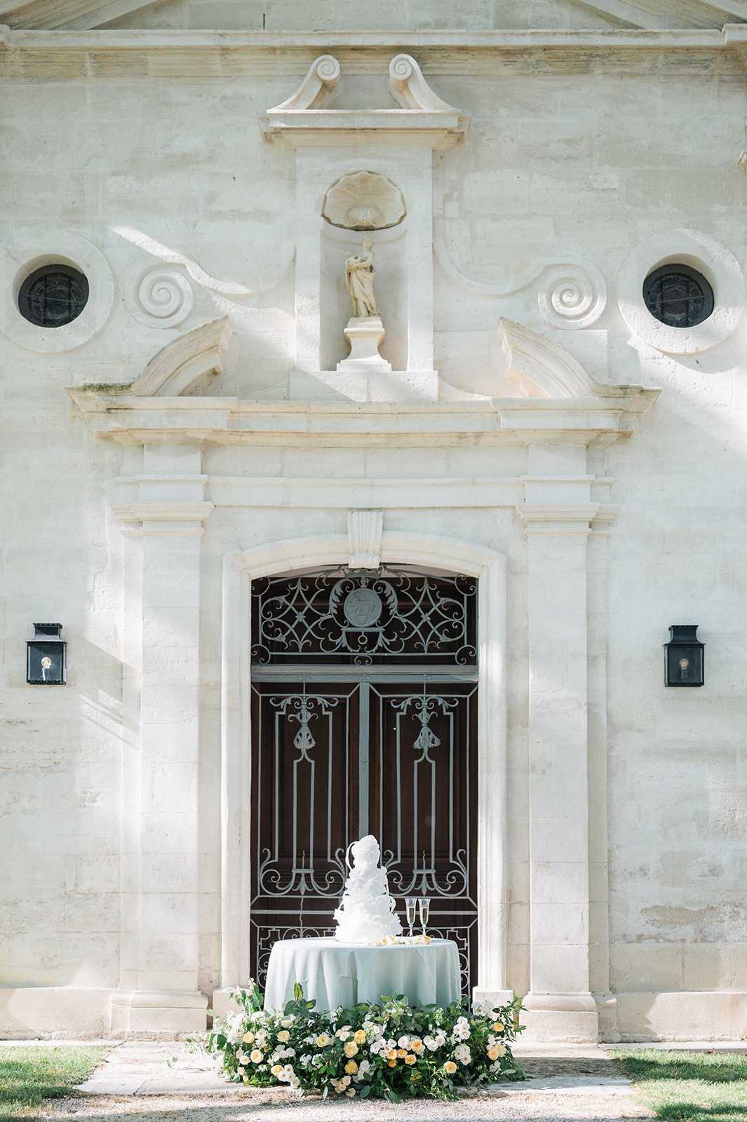 A multi-tiered white wedding cake is displayed on a small round table draped in a pale sage-green linen, positioned outdoors in front of an ornate classical French chateau entrance. The doorway features elaborate wrought-iron gates, carved stone pilasters, a decorative pediment, and a classical figure sculpture in a niche above. Two champagne flutes are placed beside the cake on the table. A lush ground-level floral arrangement of soft yellow and ivory garden roses with trailing greenery surrounds the base of the table. The composition is a wide, centered portrait shot that frames the cake against the full architectural doorway. Potential venue feature image.