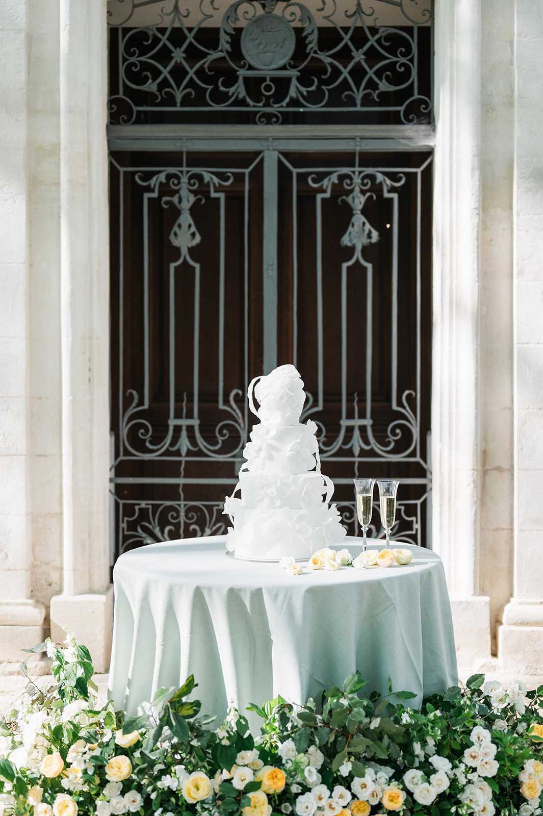 A wedding cake detail shot taken outdoors in front of a grand carved wooden door with ornate wrought-iron ironwork and stone pilasters, consistent with a French chateau or historic estate. A four-tier all-white wedding cake decorated with ruffled sugar petal appliqués is displayed on a round table draped in a pale sage green linen, accompanied by two champagne flutes filled with sparkling wine and scattered ivory rose petals. The base of the table is surrounded by a lush ground-level floral arrangement of white and soft yellow roses, white spray roses, and trailing greenery including eucalyptus. The composition is a medium shot centered on the cake table with the architectural backdrop filling the frame.