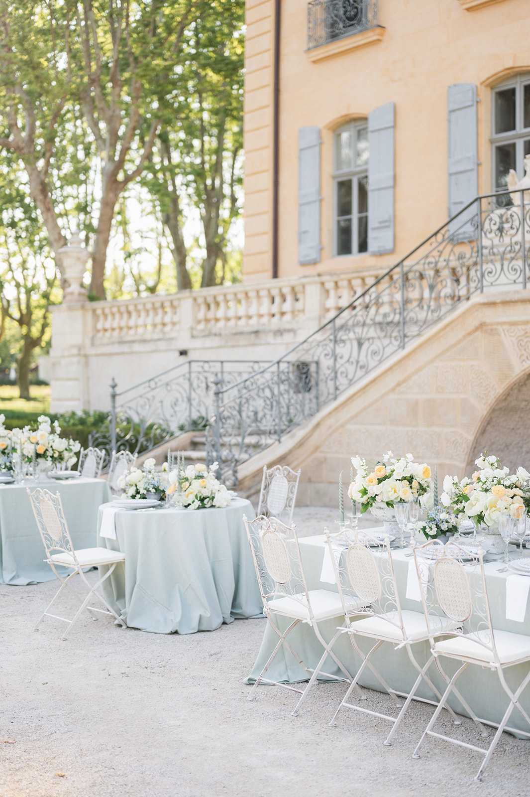 An outdoor wedding reception setup photographed in a wide shot on the gravel courtyard of a French chateau, featuring warm ochre-yellow rendered walls, pale blue-grey shutters, and an ornate wrought-iron staircase with scrollwork detailing. Multiple tables are dressed in soft sage/duck-egg blue linen — a mix of round and rectangular shapes — surrounded by white painted wrought-iron folding chairs with cream cushioned seats. Each table carries low, lush floral centerpieces composed of ivory and soft peach garden roses, white blooms, and touches of dusty blue flowers with green foliage. Place settings include grey charger plates and clear glassware. The overall decor palette is pale blue, white, and soft peach in a classic French garden style with no guests present. Potential venue feature image.