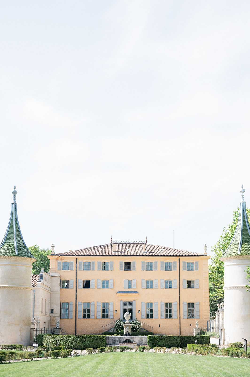 A wide exterior shot of a French chateau with warm ochre-yellow rendered walls, pale blue-grey shutters, and a terracotta tile roof. The main building is flanked by two ornate white pavilions with distinctive green pointed turret roofs topped with stone finials. A formal garden forecourt features a stone fountain urn centerpiece, neatly clipped box hedging, and a manicured lawn in the foreground. The composition is symmetrical, shot straight-on from the grounds with no people visible. Potential venue feature image.