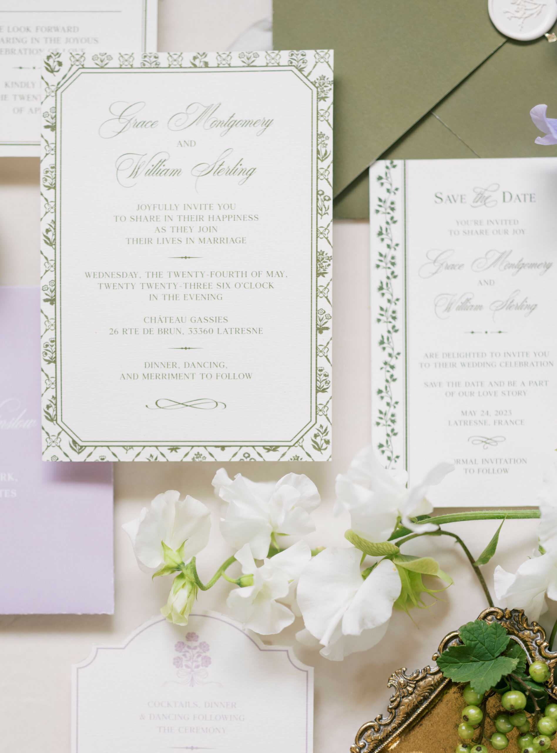 A flat lay detail shot of a wedding stationery suite for Grace Montgomery and William Sterling, with the event set for May 24, 2023 at Château Gassies, Latresne, France. The suite includes a formal invitation with a sage green botanical border on white card stock, a matching save-the-date card with a vine border, a lavender card, an olive green envelope, and a wax seal visible in the upper corner. The color palette across the suite is sage green, soft lavender, and ivory. Fresh white sweet peas are scattered across the lower portion of the flat lay, and a small ornate gold tray holding green grapes sits in the lower right corner, adding to the garden-inspired, classic French styling of the stationery design.