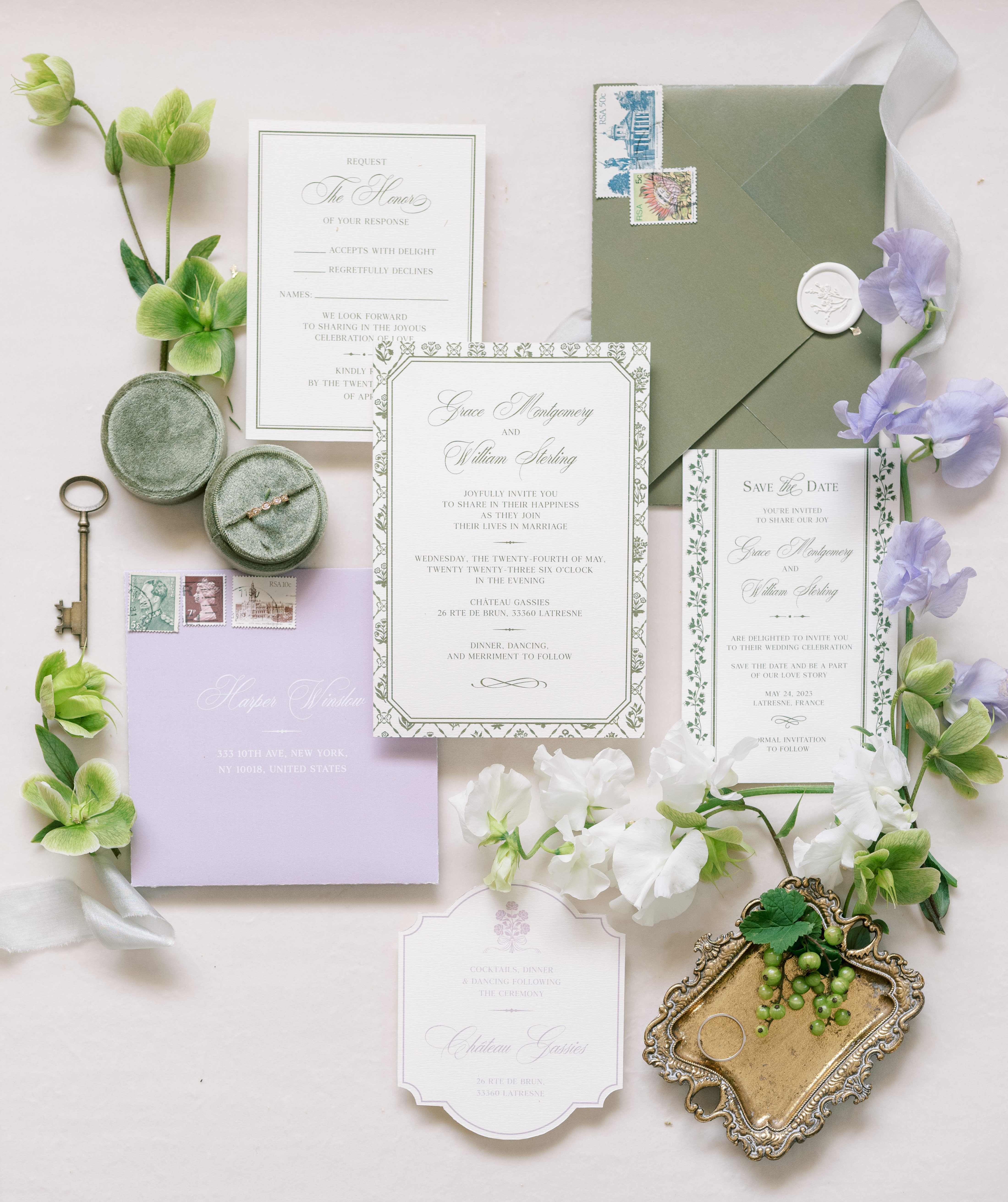 A flat lay detail shot of a complete wedding stationery suite for Grace Montgomery and William Sterling, set against a cream background. The suite includes a main invitation with a sage green botanical border, a save the date card with matching green floral detailing, an RSVP card, a sage green envelope sealed with a white wax seal and decorative stamps, a soft lavender envelope addressed to a New York recipient, and an acrylic or translucent venue details card referencing Château Gassies in Latresne, France. Styling props include a sage green velvet ring box with a diamond ring, an ornate brass key, a small antique gold tray holding two wedding bands with a decorative grape cluster, loose vintage-style postage stamps, and a length of pale grey silk ribbon. Fresh flowers scattered around the arrangement include white and lavender sweet peas and green hellebore stems, reinforcing the sage green and soft lavender color palette of the stationery suite.