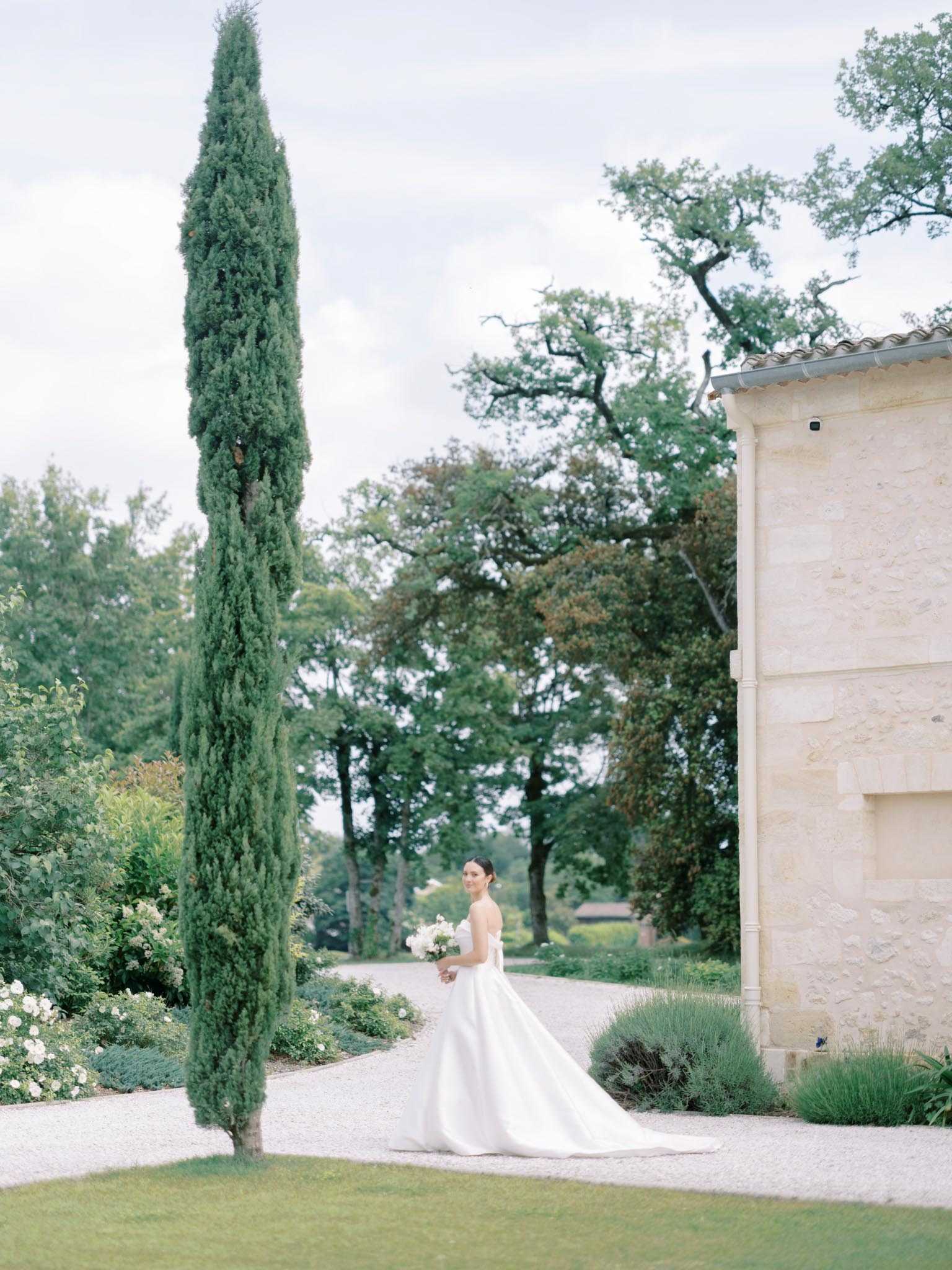 A bridal portrait taken outdoors on a gravel path alongside a stone chateau building with terracotta roof tiles. The bride stands alone, turned slightly toward the camera with a gentle expression, wearing a strapless white ball gown with a full skirt and trailing train, and holding a compact white bouquet. A tall, narrow cypress tree rises prominently in the foreground, framing the composition, while lavender plantings and white flowering shrubs border the path. The shot is a wide environmental portrait that places the bride as a mid-ground subject, emphasizing the formal garden setting and chateau architecture around her.