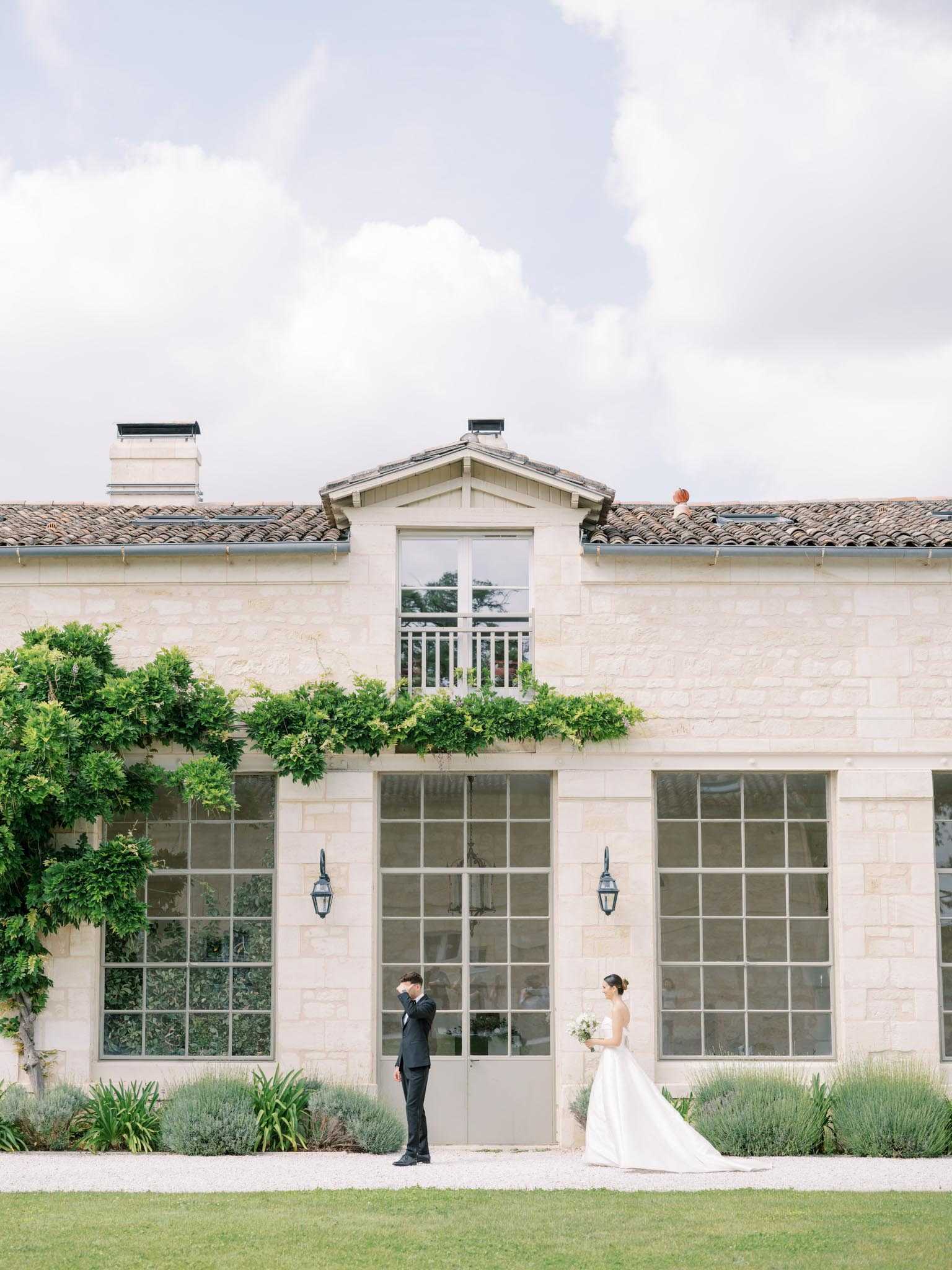 A first look moment captured outdoors in front of a classic French limestone building with large steel-framed windows, terracotta roof tiles, wall-mounted lanterns, and climbing greenery along the facade. The groom, dressed in a black tuxedo, stands with his back turned and his hand raised to his face, while the bride approaches from the right wearing a white ballgown with a full skirt and carrying a white bouquet. The two figures are positioned on either side of a central door, separated by a wide gap, suggesting the moment just before the first look reveal. The composition is a wide shot that emphasizes the building's architecture alongside the couple, with a gravel path and trimmed plantings in the foreground. Potential venue feature image.