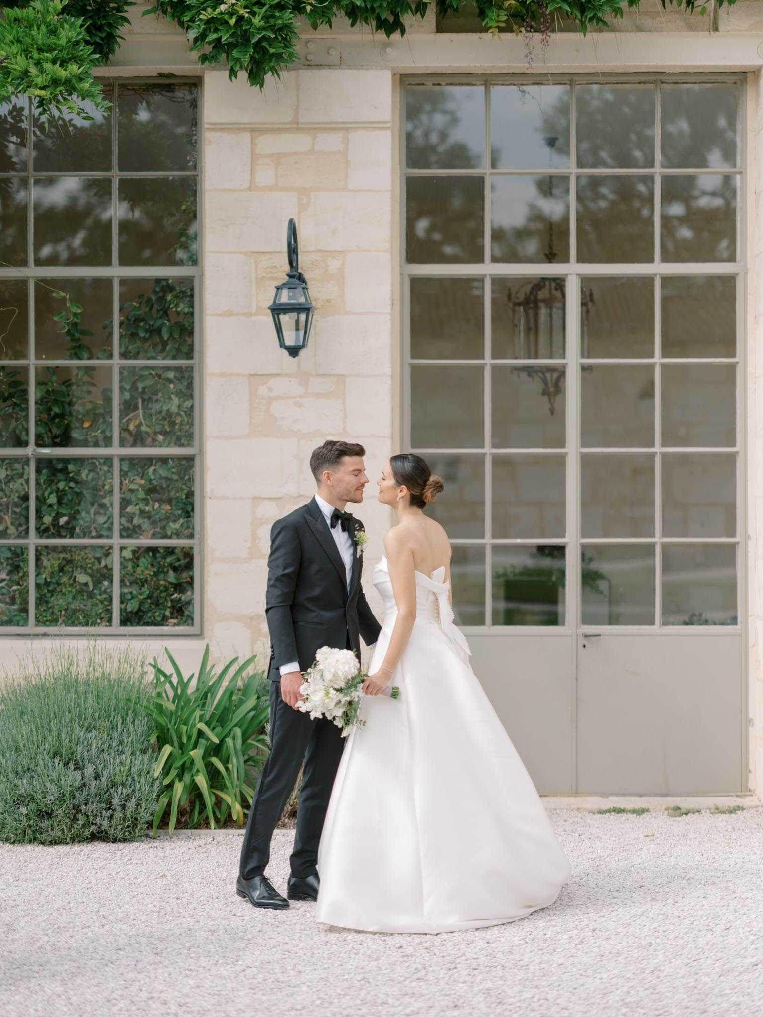 A couple portrait taken outdoors on a gravel courtyard in front of a classic French limestone building with large steel-framed windows and a black iron wall lantern. The couple is leaning in close, nearly touching foreheads. The bride wears a strapless white ballgown with a structured bow detail at the back and carries a bouquet of white hydrangeas and white blooms with minimal greenery; her dark hair is styled in a low updo. The groom wears a black tuxedo with a black bow tie and a small white boutonnière. The styling is clean and classic with a black-and-white color palette. The shot is a full-length portrait composition with the building facade centered behind the couple.