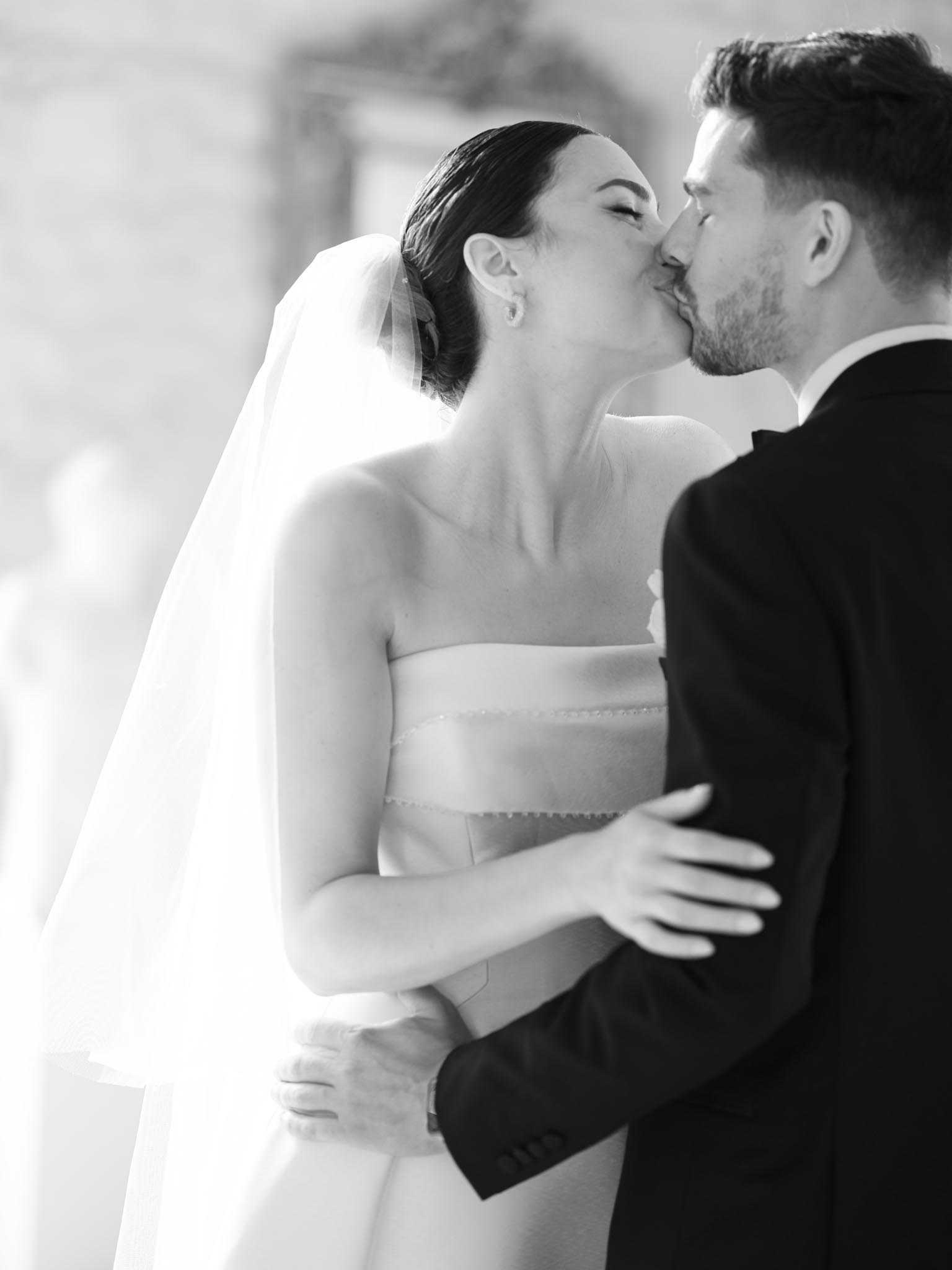 This is a black-and-white close-up portrait of a bride and groom sharing a kiss during what appears to be a couples portrait session outdoors, with softly blurred architectural elements visible in the background. The bride wears a strapless structured gown with horizontal banded detailing at the bodice, a cathedral-length veil, and small stud earrings, with her dark hair pulled back in a sleek updo. The groom is dressed in a dark suit jacket and has short dark hair with light stubble. The image is shot with a shallow depth of field, placing sharp focus on the couple's faces and upper bodies, with bright highlights and soft mid-tones giving the composition a high-contrast, polished feel.