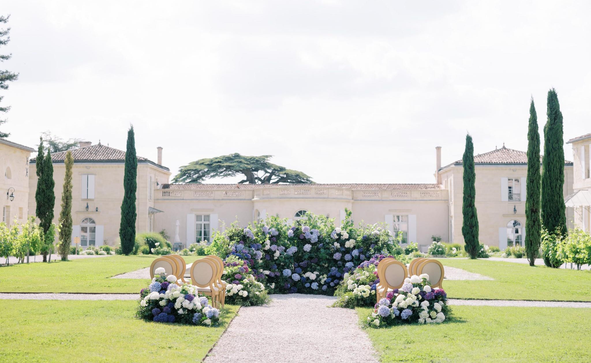 An outdoor wedding ceremony setup photographed from a wide, symmetrical angle on the grounds of a French chateau. The ceremony aisle leads to a lush floral backdrop composed of massed white, lavender, and deep purple hydrangeas interspersed with greenery, positioned at the end of a gravel pathway. On each side of the aisle's entrance, clusters of matching white and blue-purple hydrangeas flank pairs of gold-framed Louis XVI-style chairs arranged back-to-back, likely serving as ceremony seats or decorative accents. The venue features a classic limestone chateau facade with symmetrical wings, white shuttered windows, and terracotta roof tiles, with tall Italian cypress trees framing the building on both sides. The decor palette centers on white, lavender, and violet with gold chair frames, giving the setup a classic French garden style. No people are present in the image. Potential venue feature image.