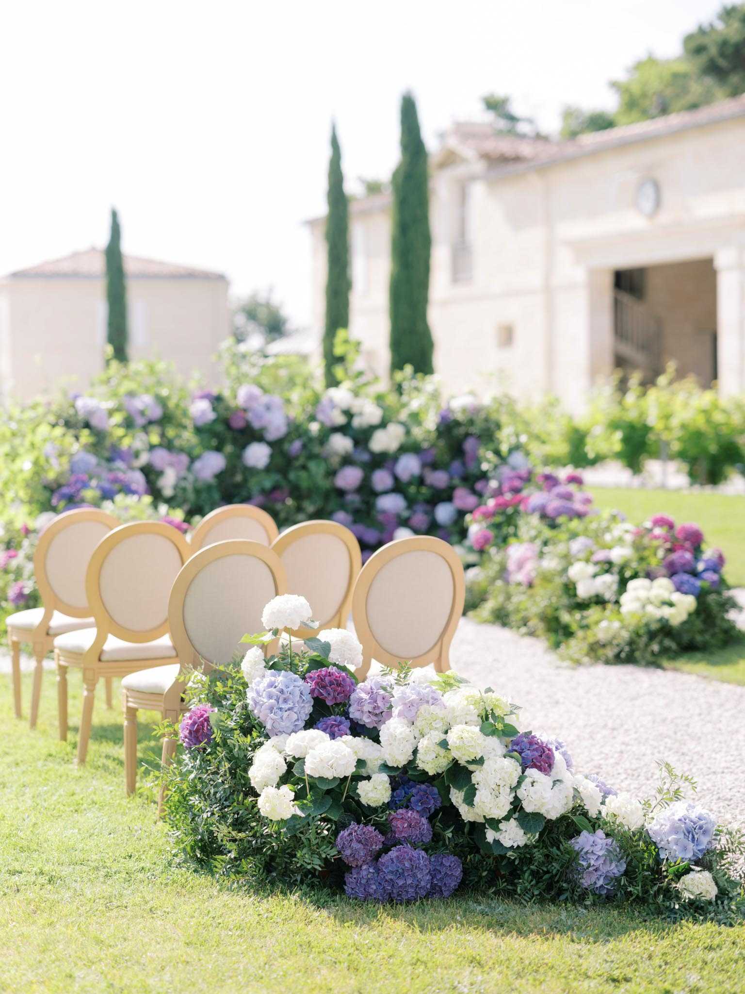 An outdoor ceremony setup on the grounds of a French stone manor, with gold-framed Louis XVI-style chairs with cream upholstery arranged in rows along a gravel aisle. The aisle is lined with large, lush arrangements of white, lavender, blue, and deep purple hydrangeas mixed with greenery, creating a garden-style floral border on both sides. The composition is a medium wide shot taken from ground level at the aisle's edge, with the chairs and floral installations in focus and the manor building softly blurred in the background. The overall decor palette is classic French with a soft purple and white color scheme. Potential venue feature image.