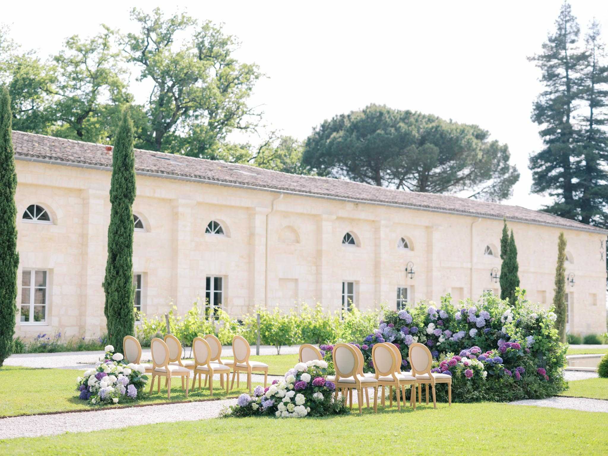 An outdoor wedding ceremony setup on a lawn in front of a large French limestone building with arched windows and a tiled roof, consistent with a chateau or domaine property. Two curved rows of gold Louis XVI-style chairs with cream upholstery are arranged in a semicircle, facing an open central aisle. The floral decor features abundant clusters of purple, lilac, and white hydrangeas arranged at ground level along the chair rows and at the ends of each row, creating a lush floral border. Tall cypress trees flank the building facade, and a vineyard is visible in the background. The wide-angle shot captures the full ceremony layout with no guests or couple present, indicating this is a pre-ceremony setup image. Potential venue feature image.