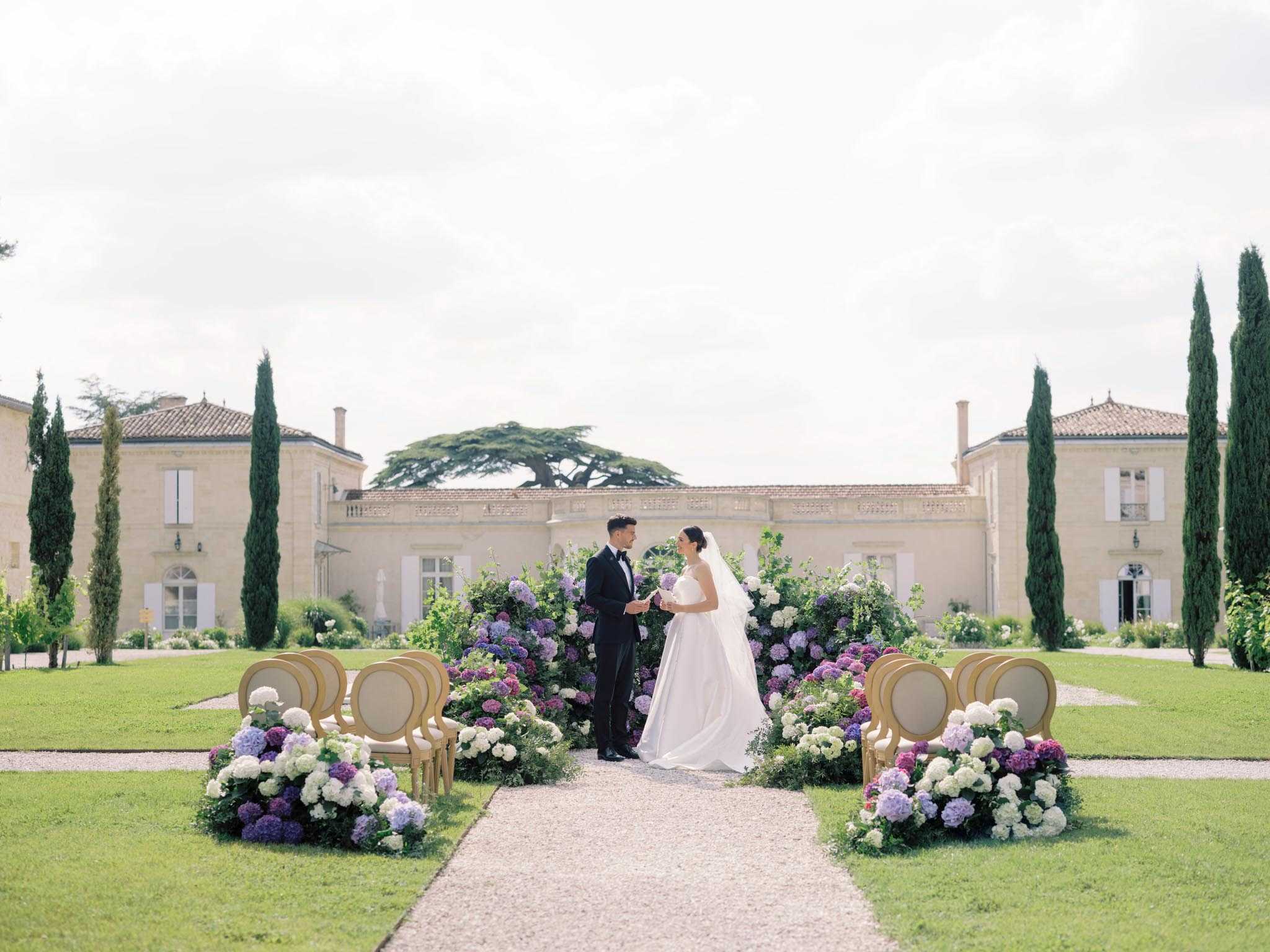 A couple stands together at the altar during an outdoor ceremony set on the formal grounds of a French château, facing each other as the groom holds the bride's hand. The bride wears a white ball gown with a long cathedral veil, and the groom is dressed in a black tuxedo with a bow tie. The ceremony setup features gold Louis XVI-style chairs arranged in rows along a gravel aisle, flanked by large ground-level floral arrangements of white, lavender, and deep purple hydrangeas with lush greenery. A dramatic floral backdrop of the same purple and white hydrangeas frames the couple at the altar. The wide shot is composed symmetrically, with the stone façade of the château centered in the background, flanked by tall Italian cypress trees on both sides. The overall decor palette is purple, white, and gold with a classic French formal garden style. Potential venue feature image.