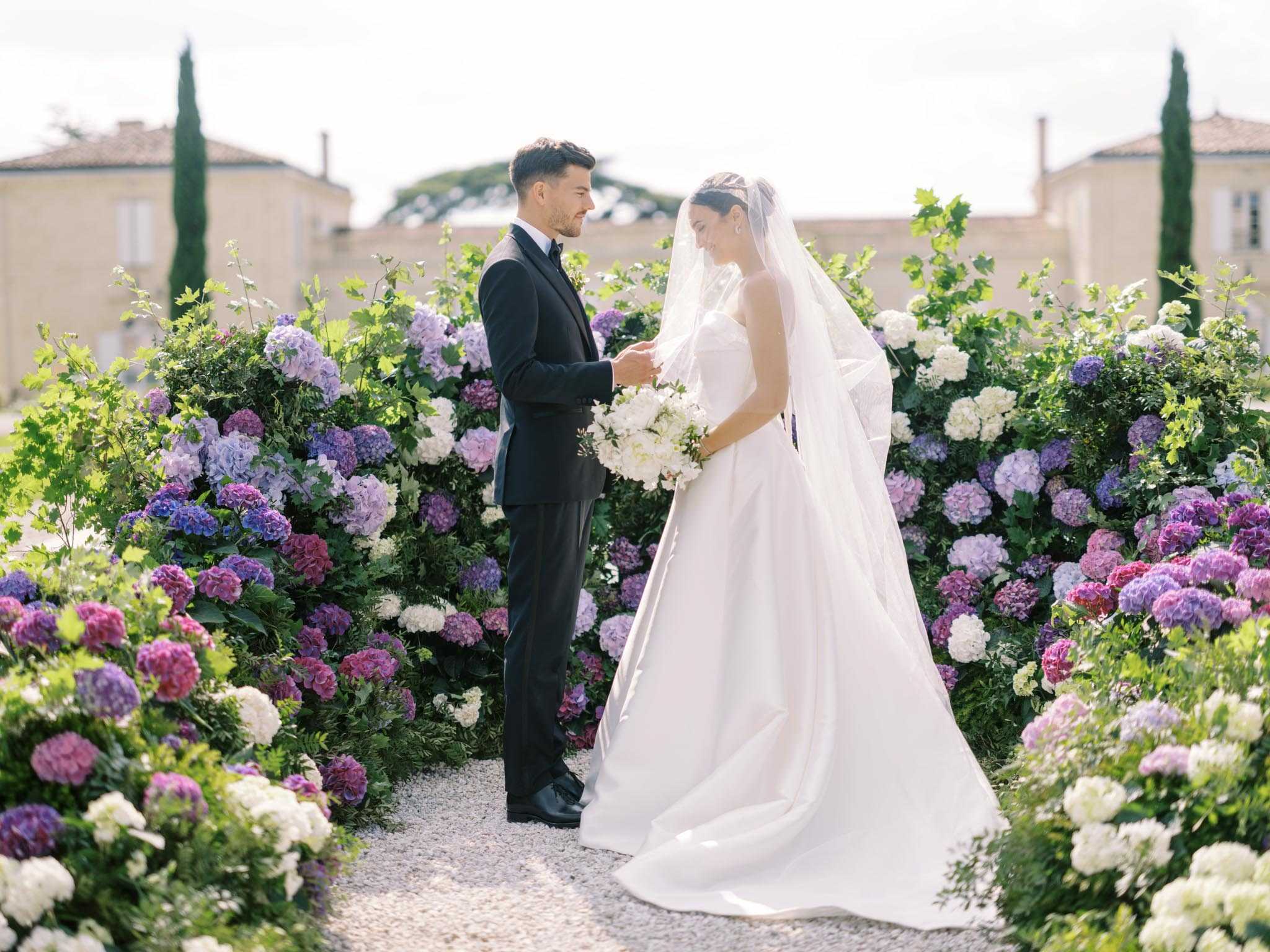 A couple portrait shot outdoors on a gravel path at what appears to be a French chateau estate, with stone buildings and cypress trees visible in the background. The bride wears a strapless ivory ball gown with a long cathedral veil and a delicate tiara, and holds a rounded bouquet of white peonies and white blooms; the groom wears a classic black tuxedo with a black bow tie. They face each other and hold hands, framed on both sides by large, lush hydrangea installations in shades of purple, lavender, magenta, pink, and white mixed with green foliage. The composition is a mid-range portrait with the floral arrangements forming a natural arch or corridor around the couple.