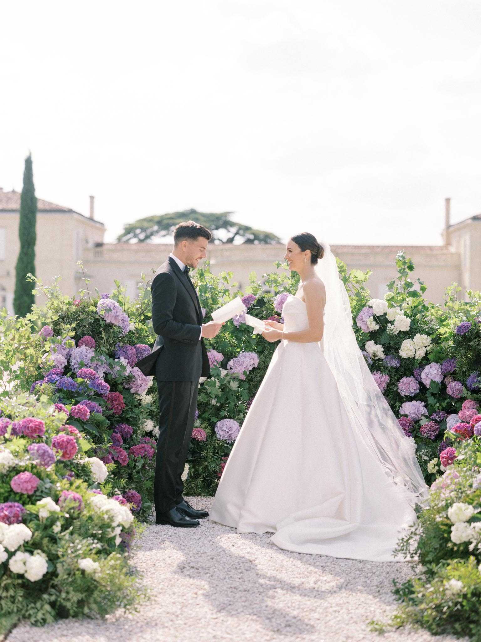 A bride and groom share a private vow reading moment outdoors on a gravel path at what appears to be a French chateau estate, with the stone building visible in the background. The groom wears a black tuxedo and the bride wears a strapless white ball gown with a full skirt and a long cathedral-length veil, her hair in an updo. They are surrounded on both sides by large, lush floral installations featuring hydrangeas in deep fuchsia, lavender, purple, and white, interspersed with green foliage, creating a corridor effect along the path. The shot is a full-length portrait taken at ground level with a slightly soft focus on the background, emphasizing the couple in the center of the frame.