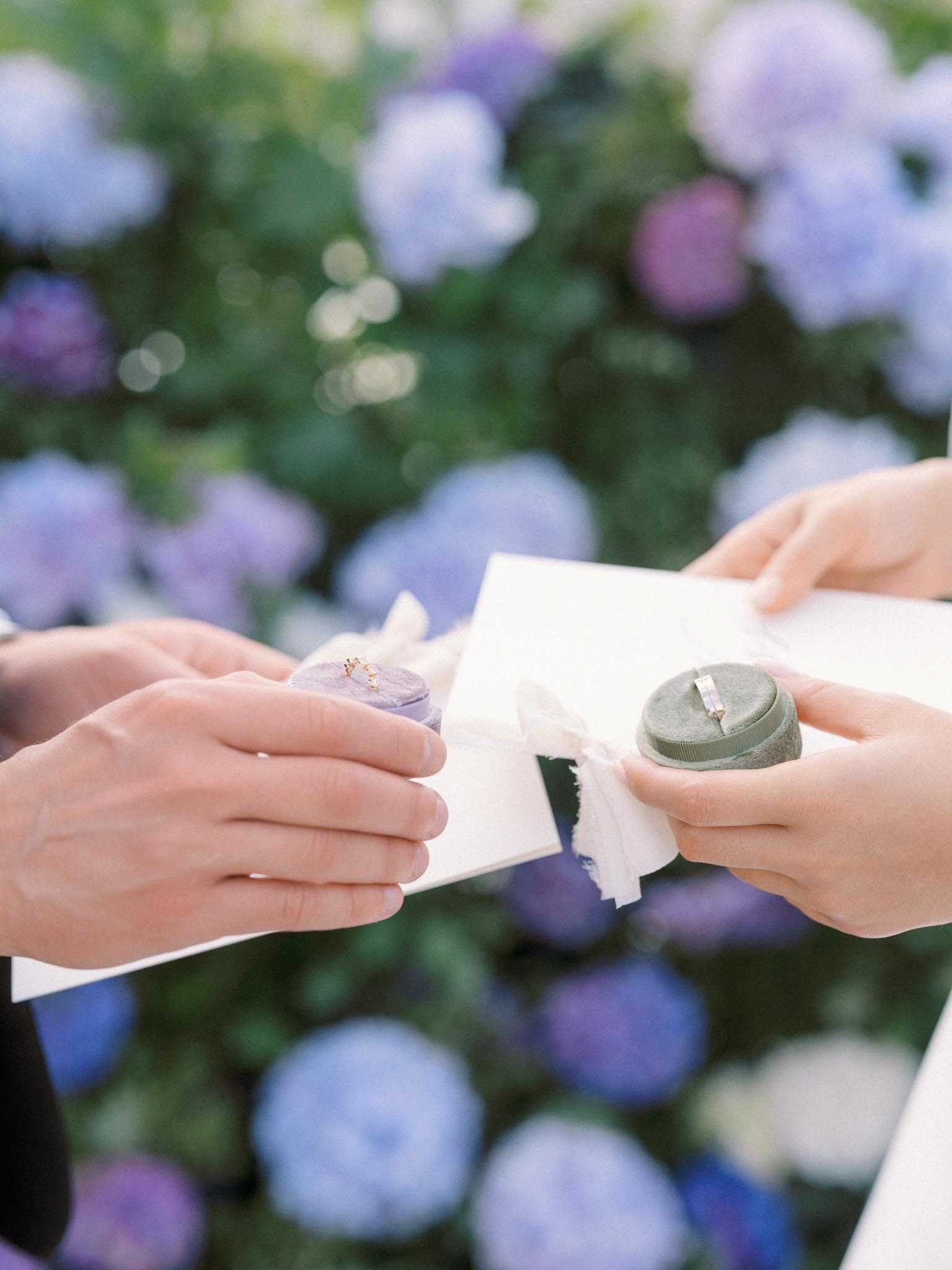 Close-up detail shot of the ring exchange moment during an outdoor ceremony, with two sets of hands holding a white deckle-edged paper card or vow booklet tied with a sheer white ribbon, and two velvet ring boxes — one lavender and one sage green — each holding a delicate gold wedding band. The background is filled with blurred blue, lavender, and purple hydrangea blooms, creating a soft, color-coordinated backdrop that echoes the tones of the ring boxes. The styling reflects a romantic, garden-inspired aesthetic with a cohesive cool-toned color palette.