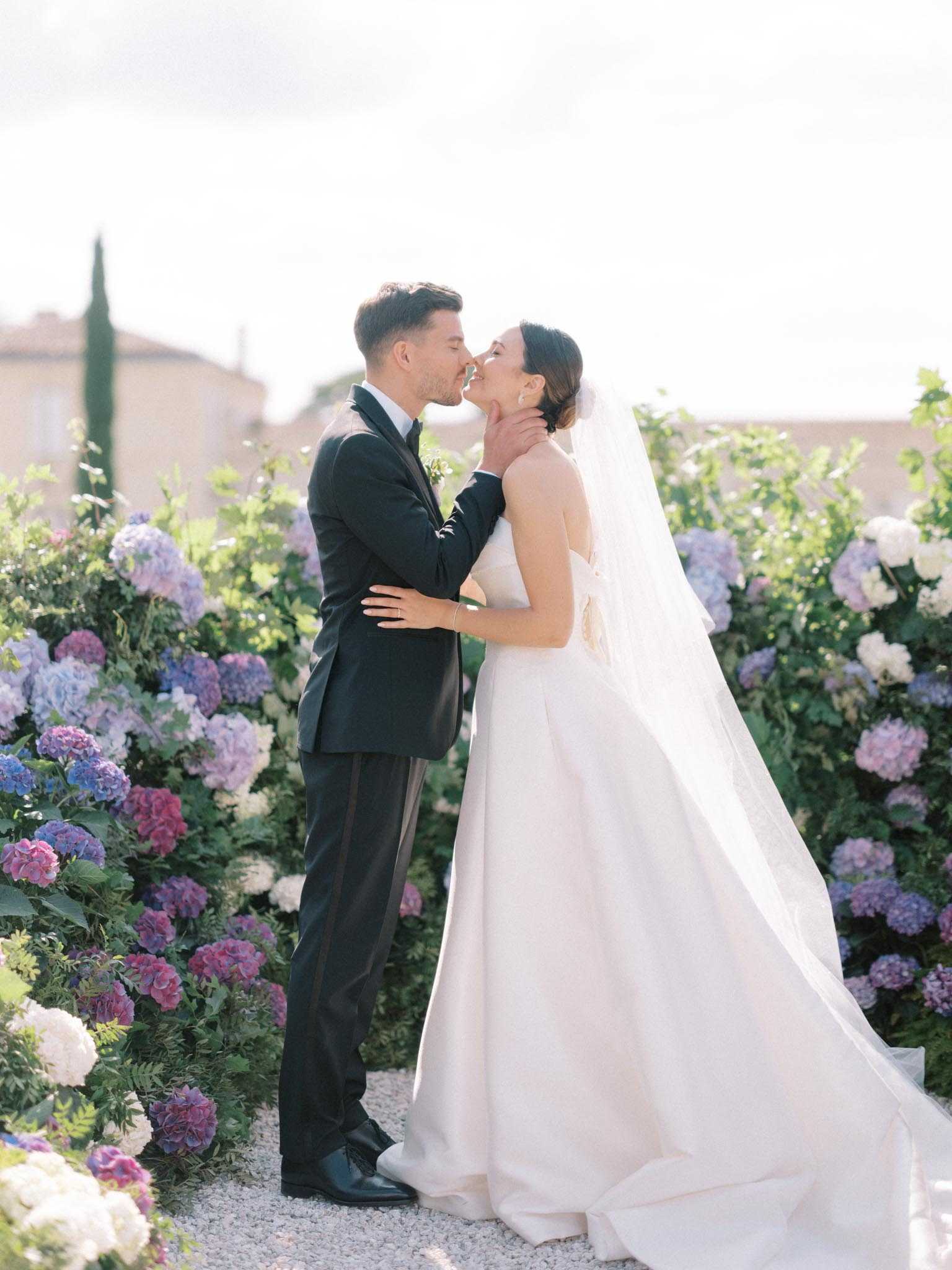A couple portrait taken outdoors in a garden setting, with the bride and groom sharing a kiss on a gravel path. The groom wears a black tuxedo with a white dress shirt and boutonniere, and gently cups the bride's face with one hand. The bride wears a strapless ivory ballgown with a full skirt and a long cathedral-length veil, with her hair styled in an updo. They are surrounded on both sides by large hydrangea bushes in shades of blue, purple, pink, and white. A stone building and tall cypress trees are visible in the soft-focus background, suggesting a Provençal or southern French estate setting. The image is a full-length portrait shot with a bright, airy exposure and soft natural light.