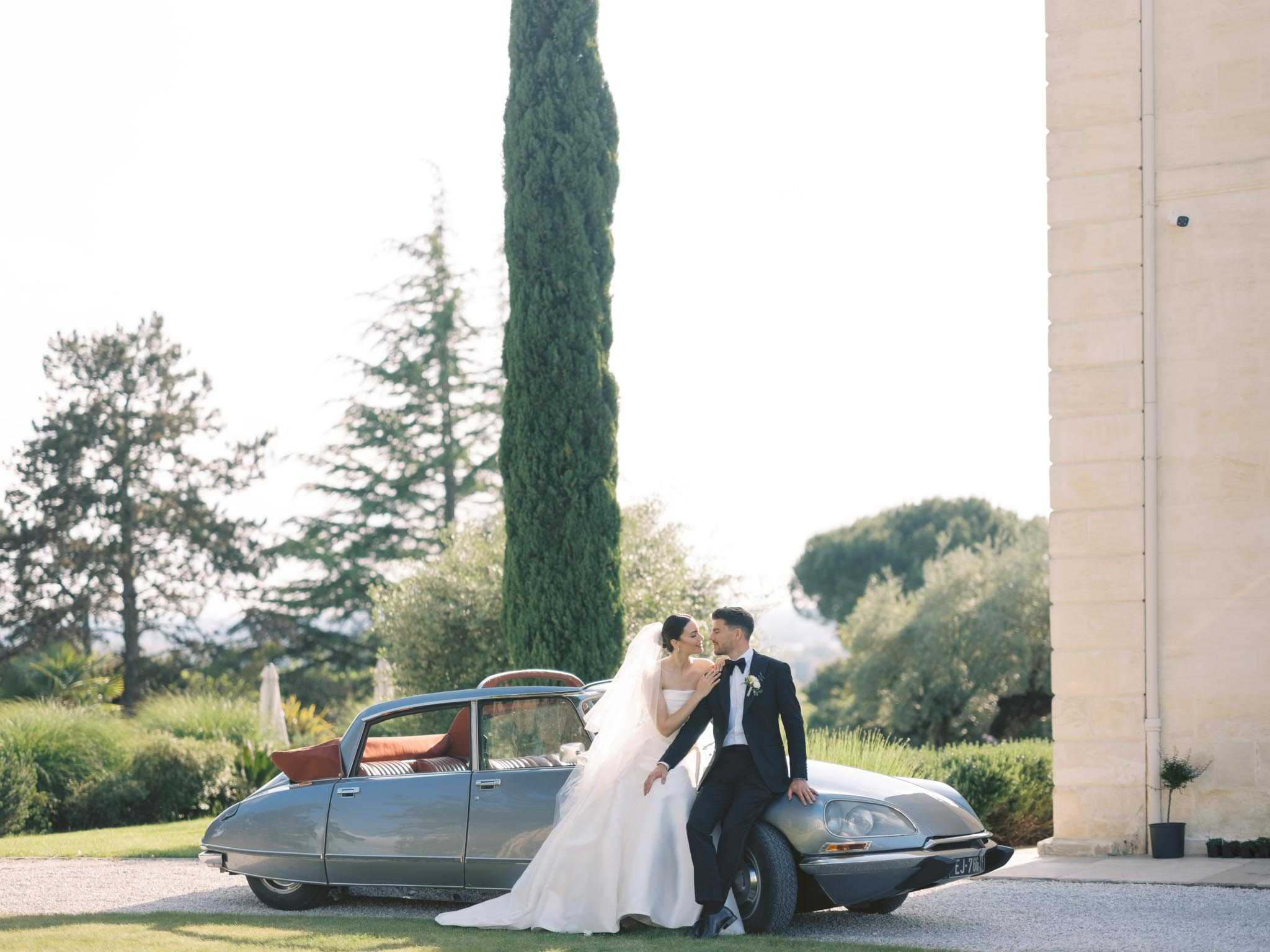 A couple portrait taken outdoors on gravel grounds beside a vintage silver Citroën DS with red interior cushions visible through the rear window. The bride wears a strapless white ballgown with a long cathedral-length veil, while the groom is dressed in a navy tuxedo with a black bow tie and a white boutonnière. The two are leaning against the front of the car, facing each other closely. The setting features the stone facade of a French chateau or estate visible on the right edge of the frame, with well-maintained grounds and landscaping in the background. The composition is a wide portrait shot taken in warm afternoon light.