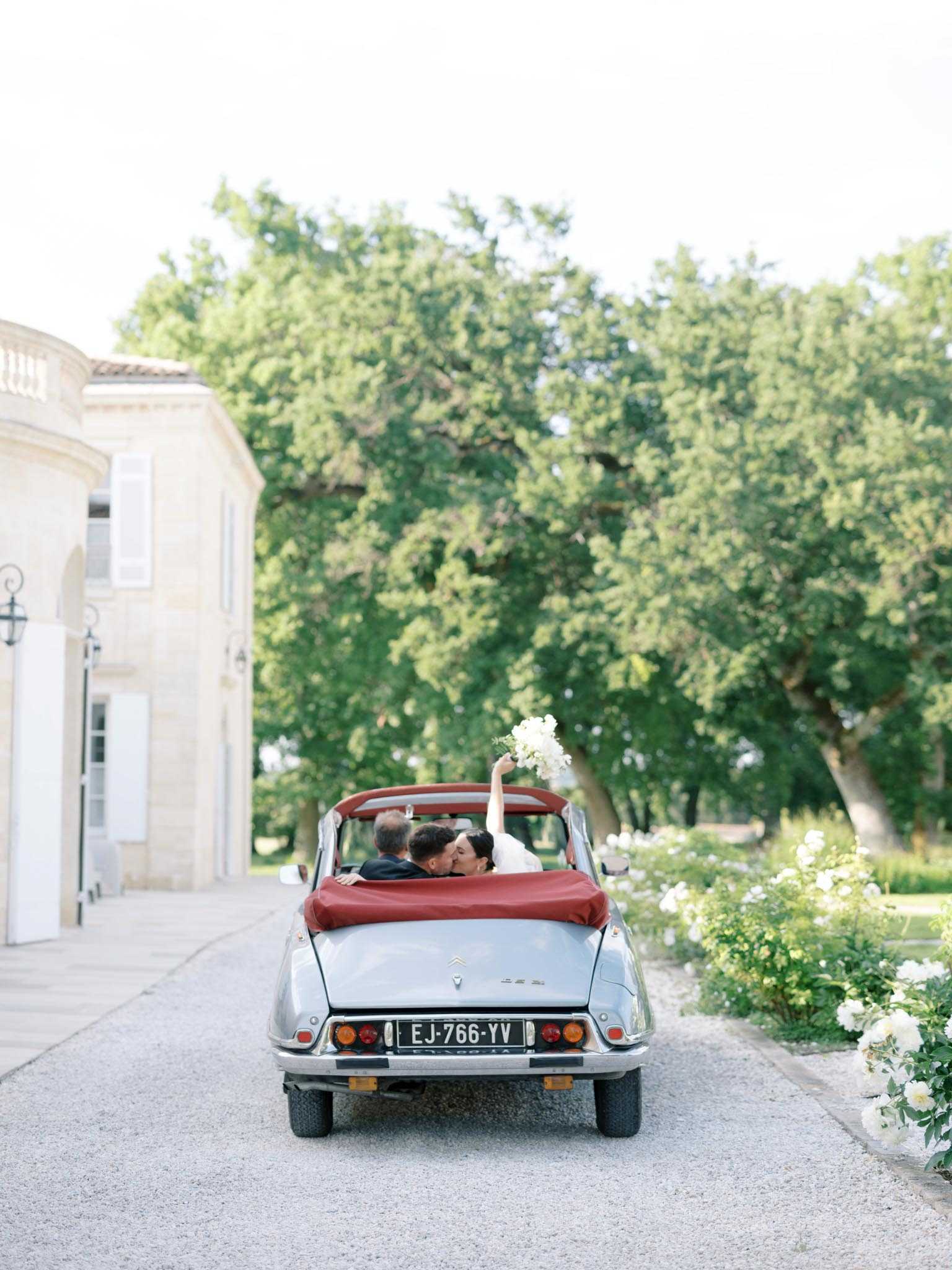 A couple shares a kiss in the back seat of a classic silver Citroën DS convertible with a red interior, photographed from behind as they depart along a gravel driveway. The bride holds a white bouquet aloft with one arm while leaning in to kiss the groom, who is dressed in a dark suit. The setting is the exterior grounds of a French chateau, with cream limestone architecture visible on the left and white flowering garden shrubs lining the right side of the drive. The shot is a wide portrait-style image taken from ground level behind the vehicle, capturing the full rear of the car and the couple framed by mature trees in the background.