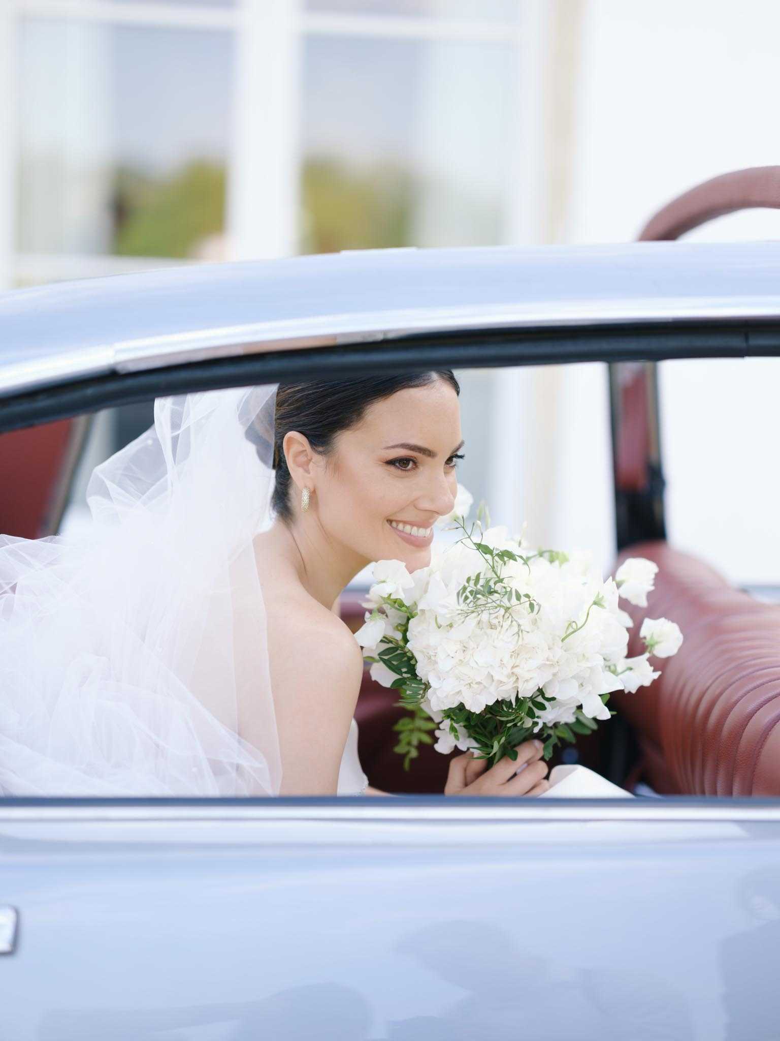 A bride is seated in the back of a vintage pale blue car with burgundy leather interior, leaning slightly toward the open window and smiling at the camera. She wears a white gown with a tulle veil, her dark hair styled in a sleek updo, and small gold drop earrings. She holds a rounded bouquet of white hydrangeas, white sweet peas, and green foliage. The shot is a close-up portrait framed by the car door and window, with a softly blurred architectural background featuring white columns.