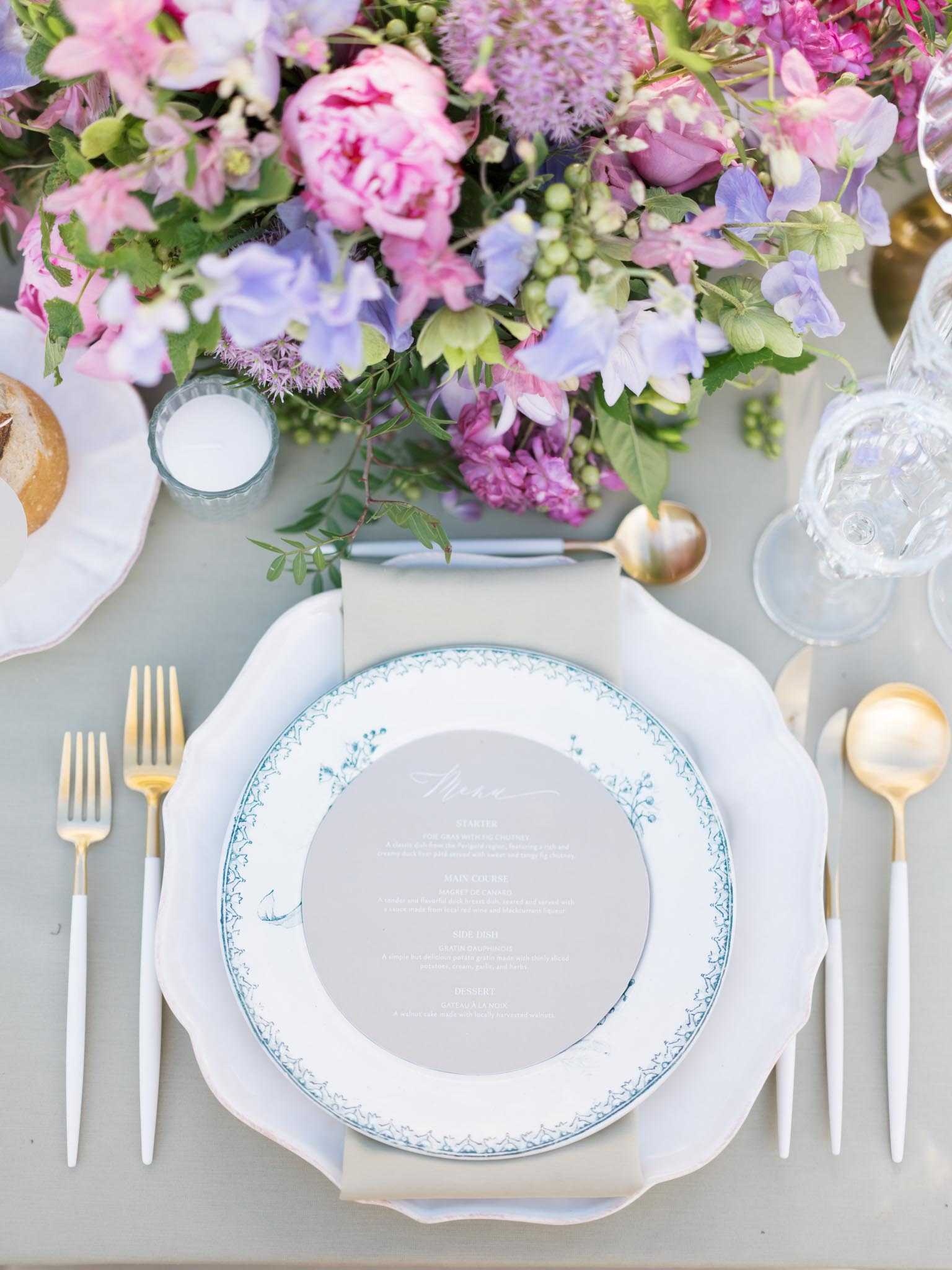 A close-up, overhead detail shot of a single place setting at a wedding reception table. The place setting layers a white scallop-edged charger plate, a white dinner plate with a delicate teal blue floral border pattern, and a circular grey menu card with white calligraphy script listing starter, main course, side dish, and dessert courses. Cutlery is gold with white handles, including two forks on the left and a knife and spoon on the right. A taupe linen napkin is folded beneath the menu card, and a small white votive candle sits nearby. The centerpiece arrangement, partially visible in the upper portion of the frame, features a lush mix of hot pink peonies, lavender sweet peas, purple allium, magenta stocks, and trailing greenery in a pink and purple palette. A clear glass tumbler and gold-toned serving spoon are visible at the upper right. The overall table decor palette combines sage green, soft grey, white, and gold with the vivid floral colors as a contrast.