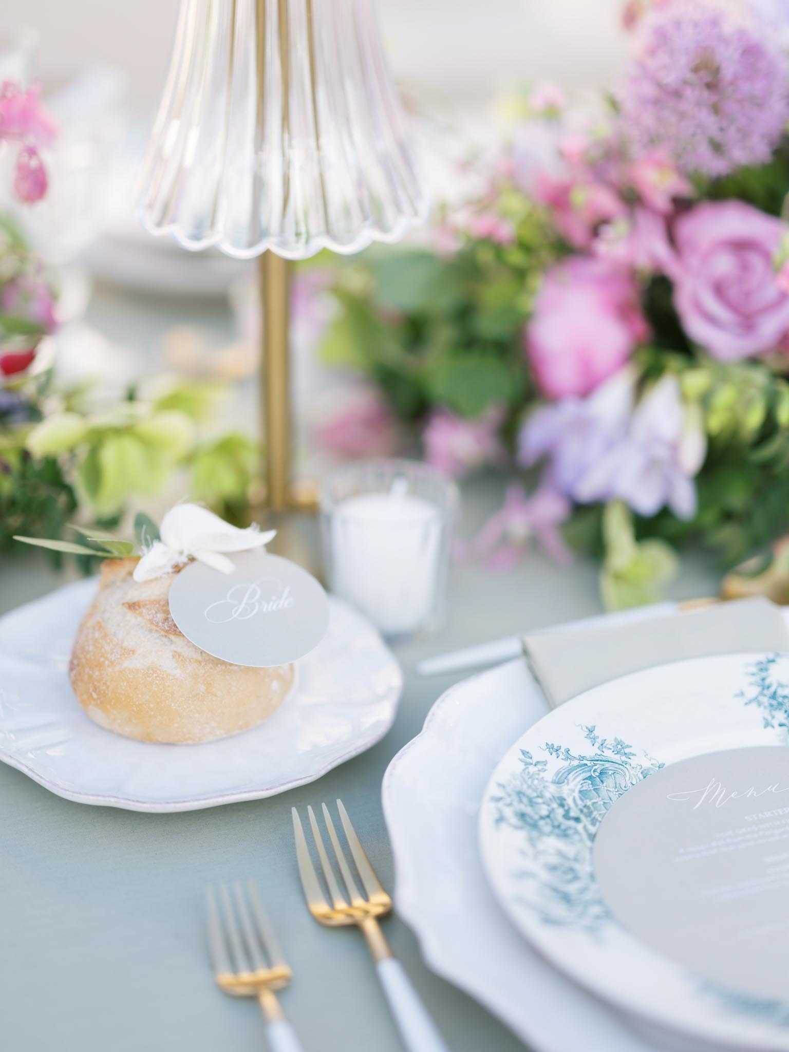 A close-up detail shot of a wedding reception table setting, specifically the bride's place setting. The place setting features white scallop-edged china with a blue toile floral pattern on the charger plate, layered with a plain white dinner plate, and a grey linen napkin. Gold flatware with grey handles is arranged to the left of the plates. A small individual bread roll sits on a separate white scallop-edged side plate, topped with a pale grey oval calligraphy place card reading 'Bride' and a small white floral accent. A small white votive candle sits nearby, and a gold table lamp with a ribbed white scalloped shade is partially visible above. The table linen is a muted sage or dusty blue-grey. Soft-focus florals in the background include pink garden roses, lavender hydrangeas, and lilac sweet peas in a lush centerpiece arrangement. The overall decor palette combines dusty blue-grey, gold, and soft pink and lavender tones in a classic French-inspired style.