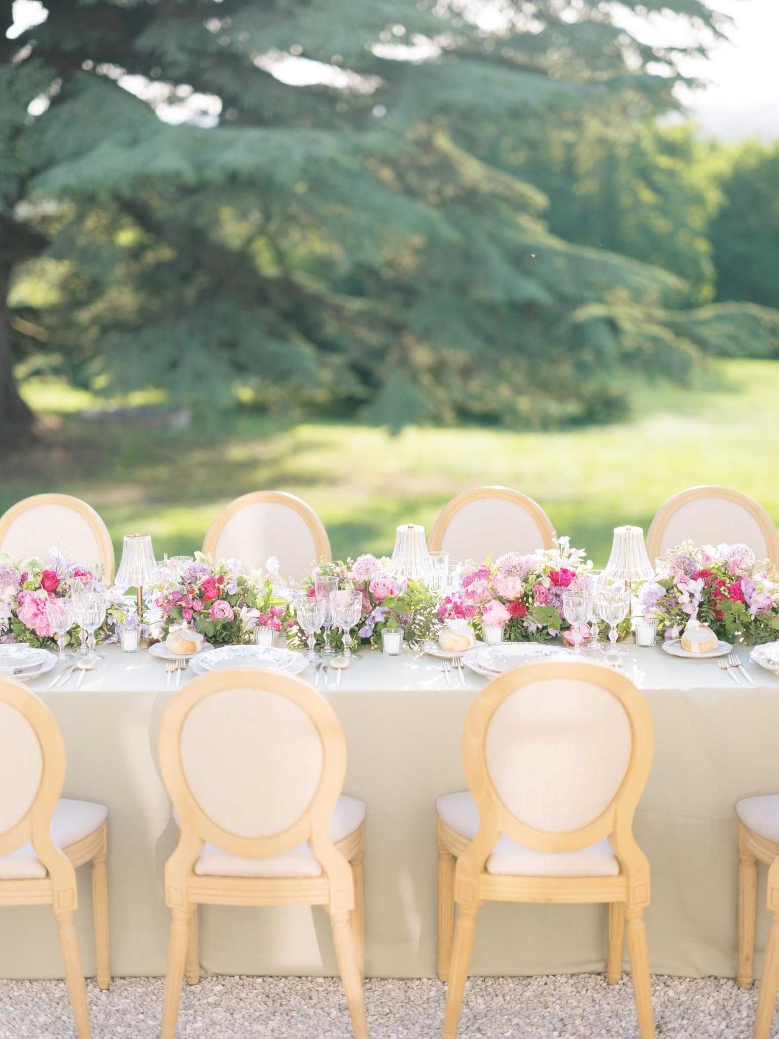 A detail shot of an outdoor wedding reception table set on a gravel surface, with no guests present. The long rectangular table is dressed in a pale ivory linen and lined with natural wood Louis XVI-style medallion chairs upholstered in cream. A lush floral runner runs the length of the table, composed of hot pink roses, blush and mauve ranunculus, lavender sweet peas, alliums, and mixed greenery. Crystal glassware, white charger plates with decorative rims, and folded napkins with bread rolls are set at each place, alongside slender glass candle holders. The overall decor palette combines ivory, blush, lavender, and fuchsia in a classic French garden style.
